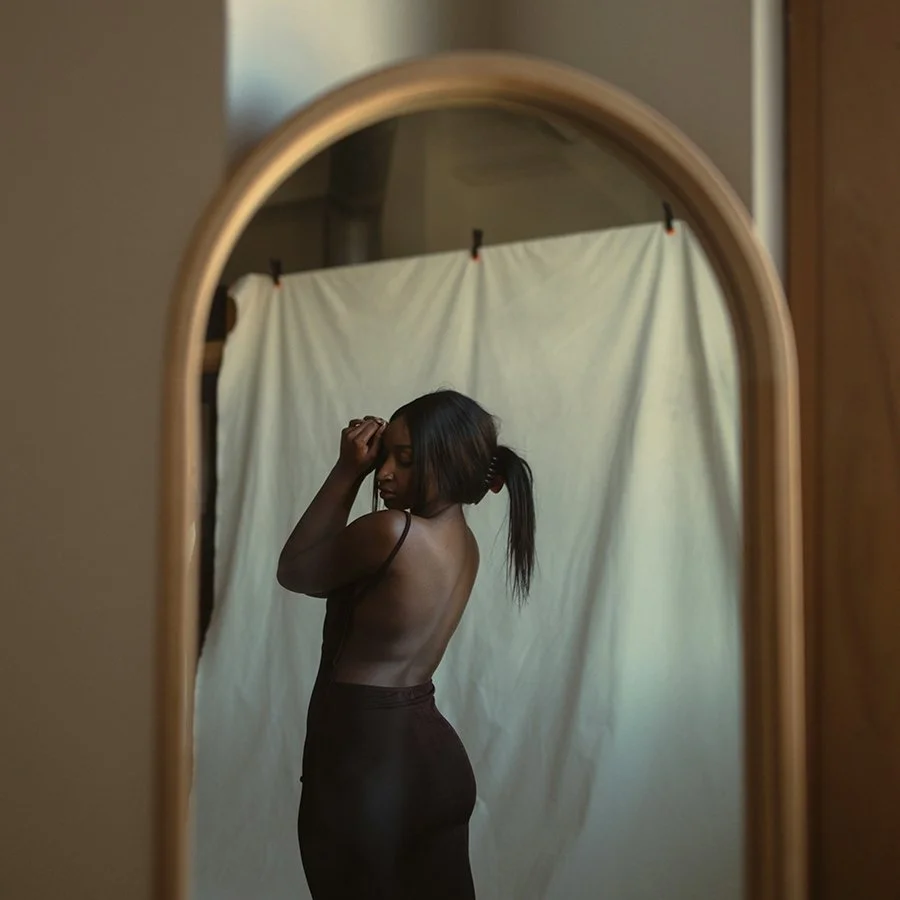 A woman standing in front of a mirror, posing with one hand on her head and wearing a black tank top and dark pants, with a light-colored curtain as the background.