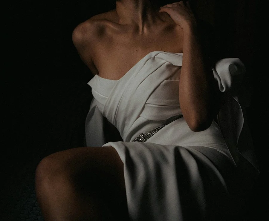 A person draped in white fabric, sitting with one arm resting on their shoulder, in a low-light setting.