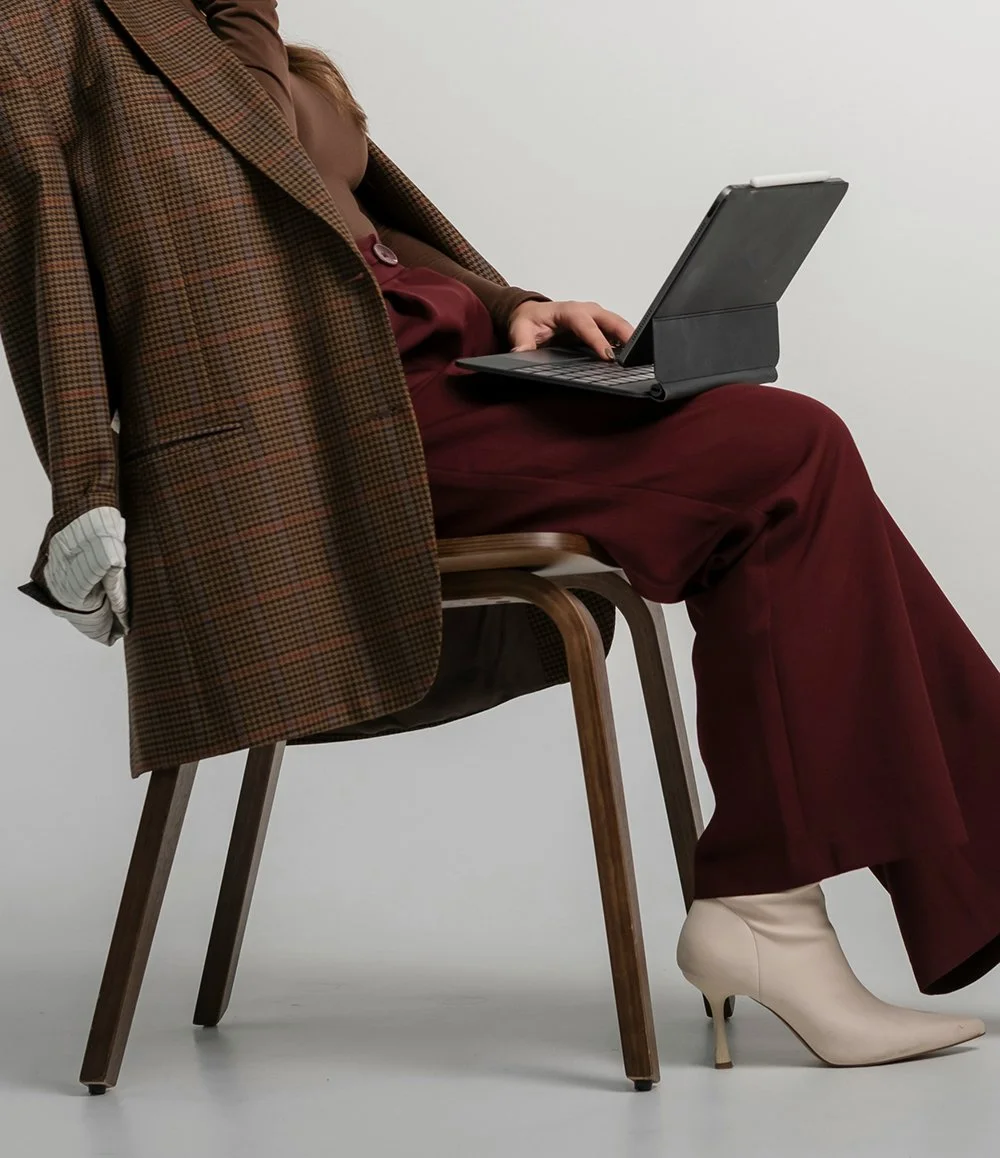 Person sitting on a wooden chair with crossed legs, wearing a brown checkered blazer, burgundy pants, beige heeled boots, and using a black tablet.