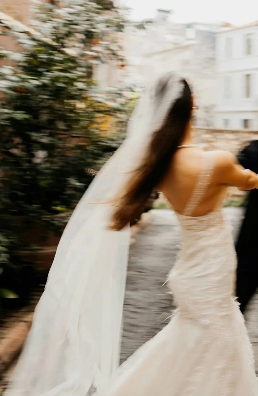 A woman in a white lace wedding dress with a long train walking outdoors, with blurred greenery and buildings in the background.