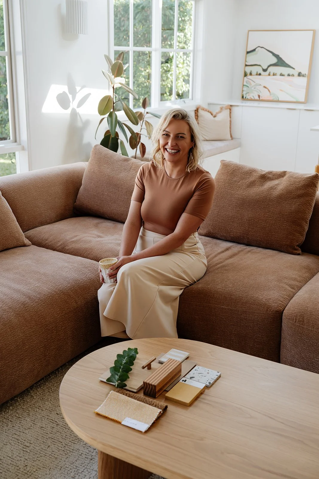A woman sitting on a beige sofa in a bright living room, holding a mug, smiling, with large windows and artwork in the background, and a wooden coffee table with decor in front.