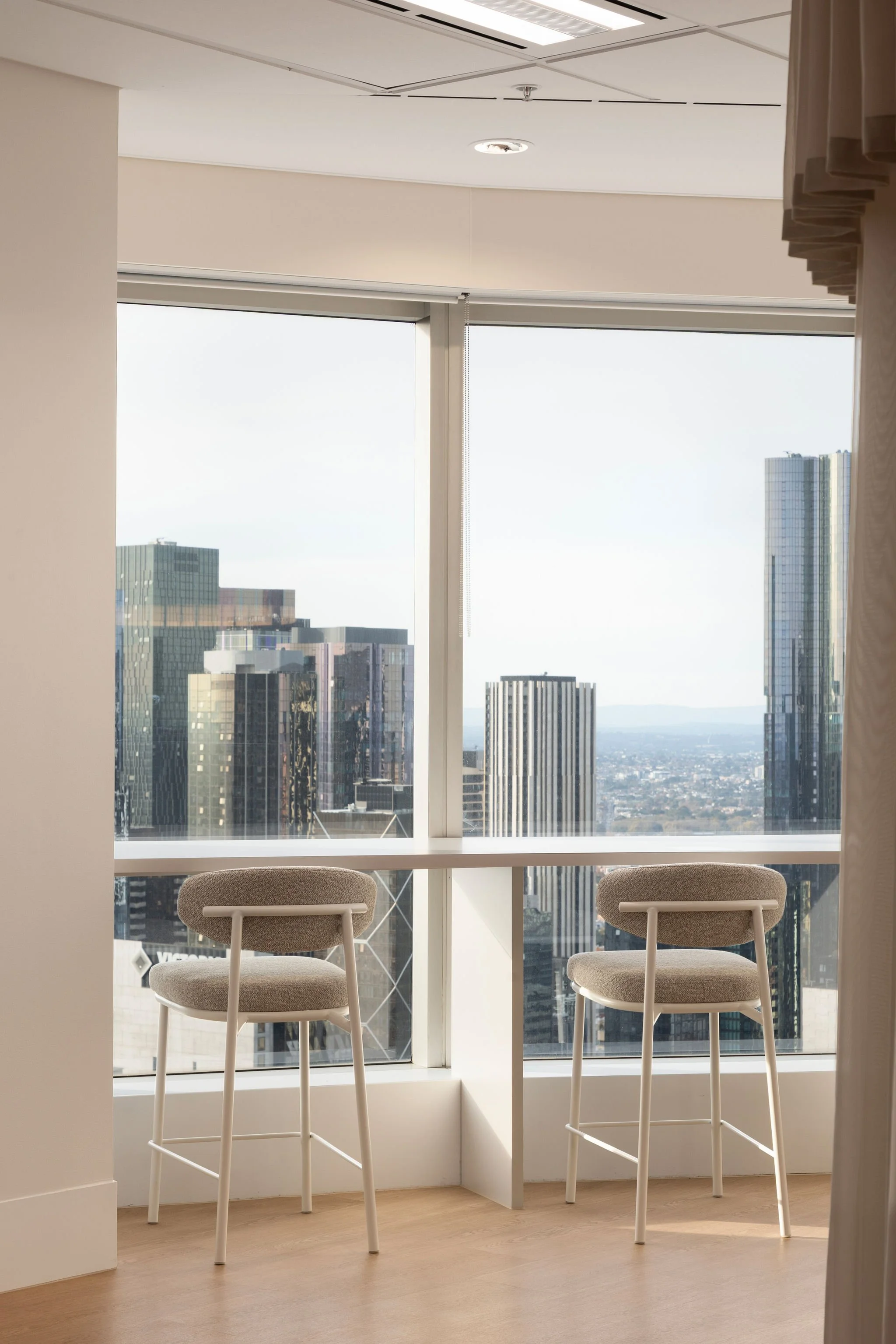 Interior of a high-rise office with three beige chairs facing a large window. The window offers a view of city skyscrapers and a distant horizon. The ceiling has built-in lighting and an air vent.