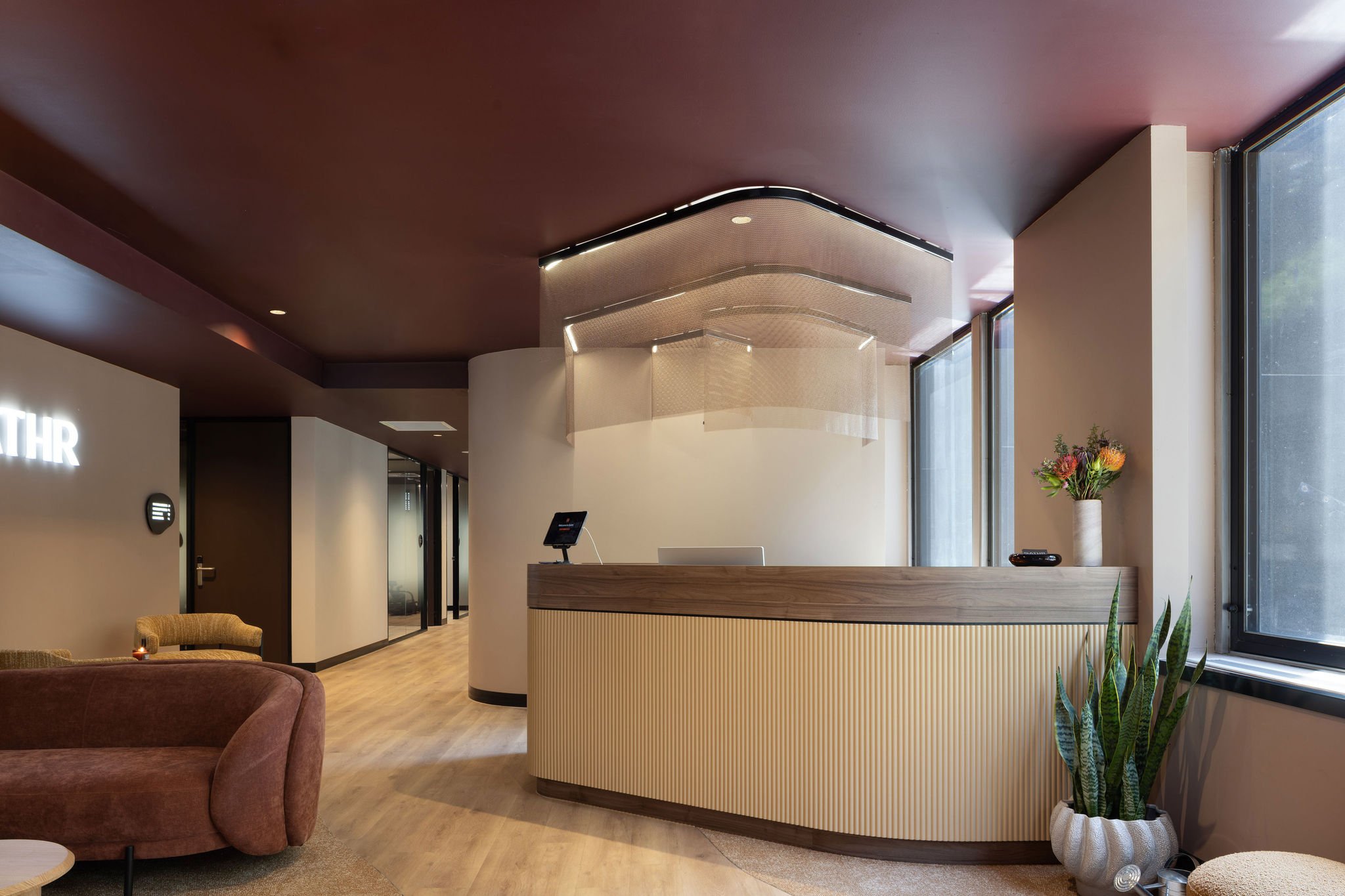 Modern reception area with a curved beige and black front desk, large windows on the right, a tall flower vase on the right side, a partial view of a brown armchair on the left, and a lounge area with beige seats on the left. The ceiling is dark purple with integrated lighting, and there's a decorative hanging ceiling feature.