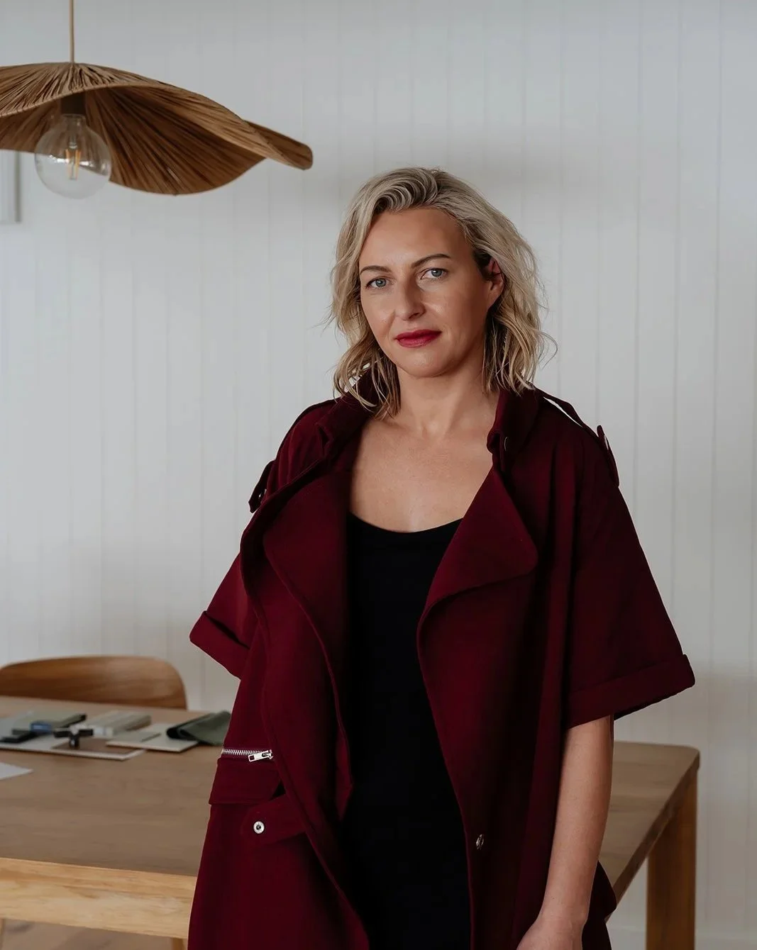 A woman with blonde wavy hair wearing a burgundy jacket and black top stands in a room with beige wooden furniture and a modern wooden ceiling light.