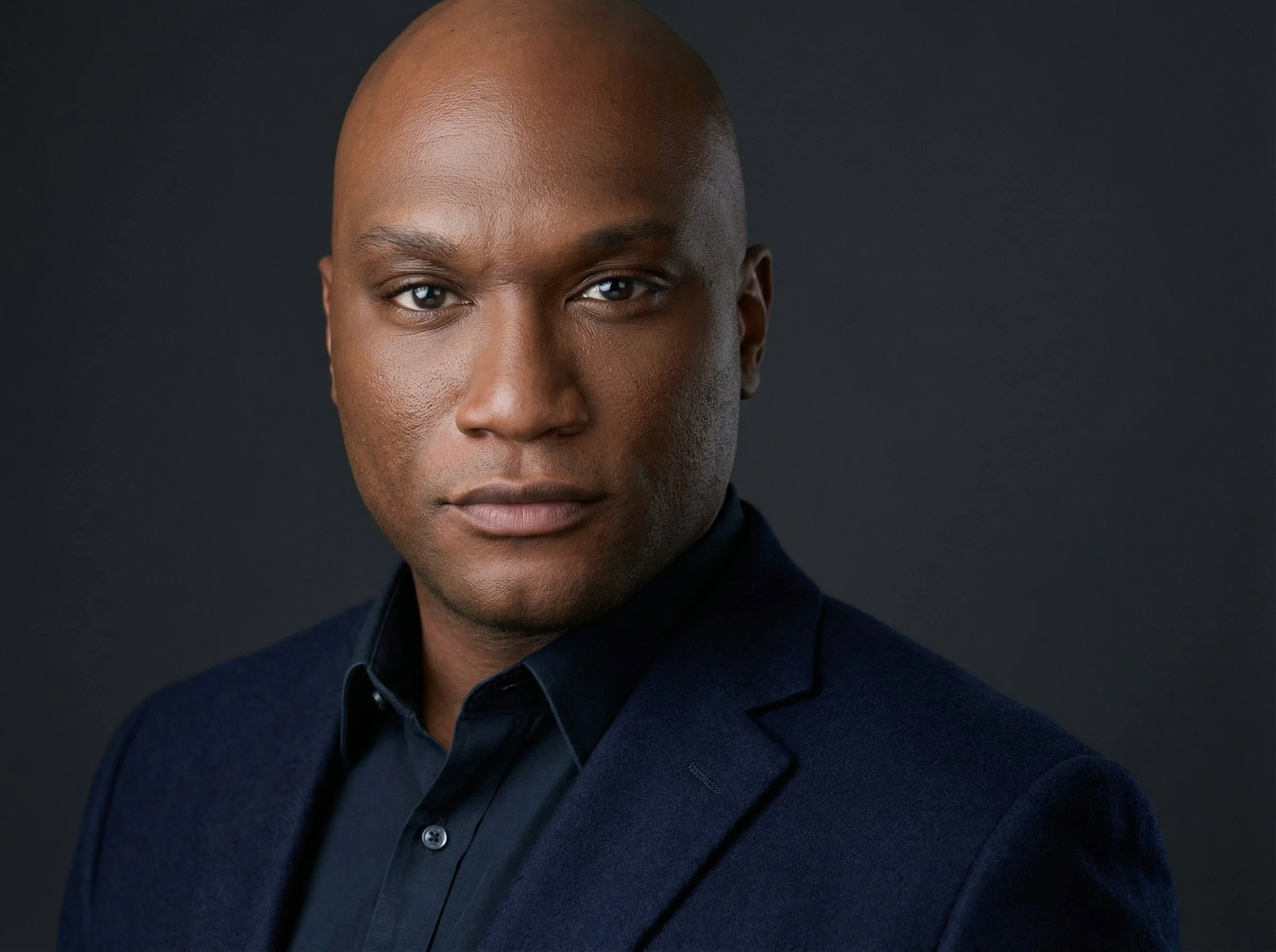 Close-up portrait of a confident Black man with a smooth bald head, wearing a dark blazer and shirt against a dark background.