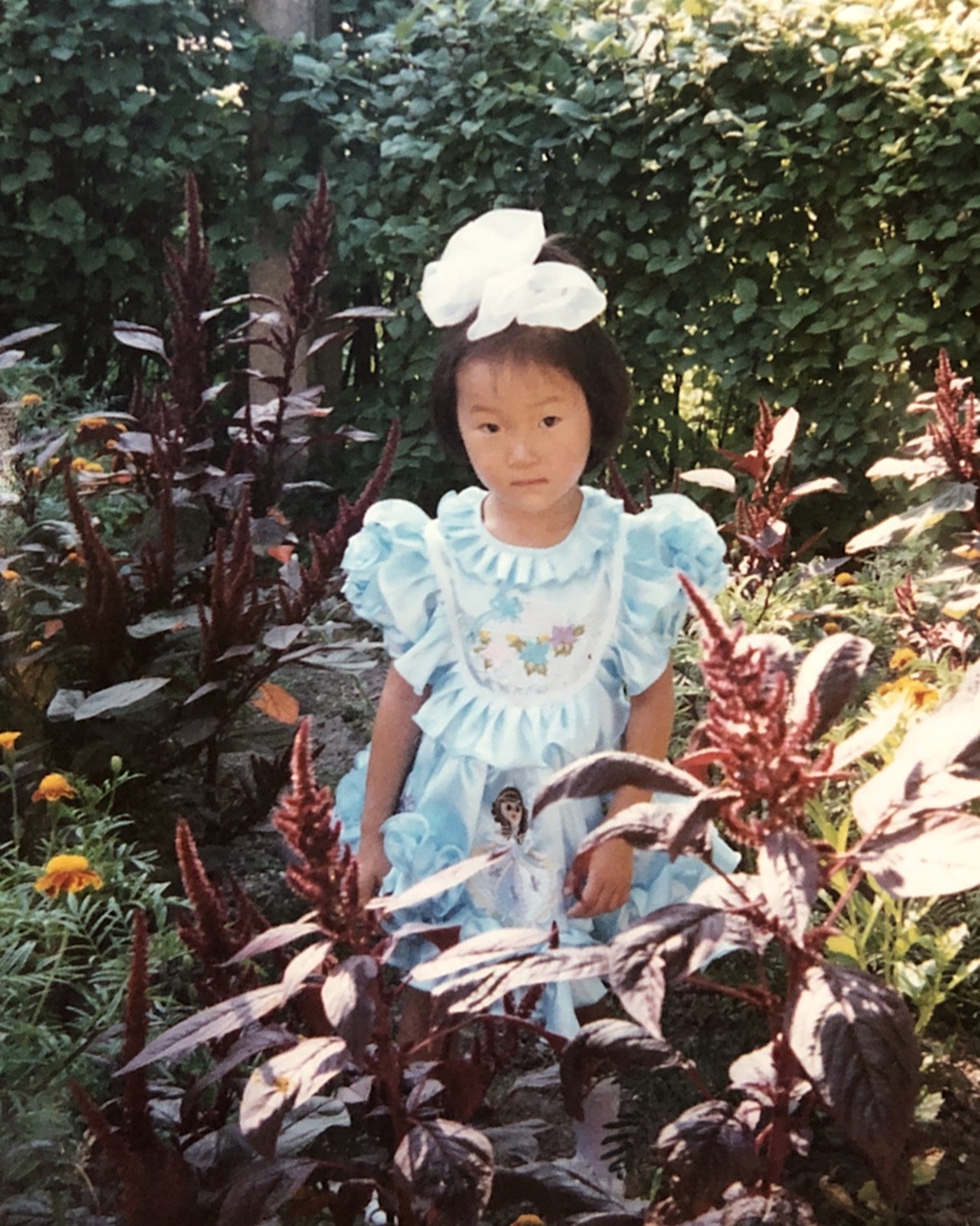 Marina Ogai as a little girl in a pale blue dress and a big white bow, standing among garden flowers