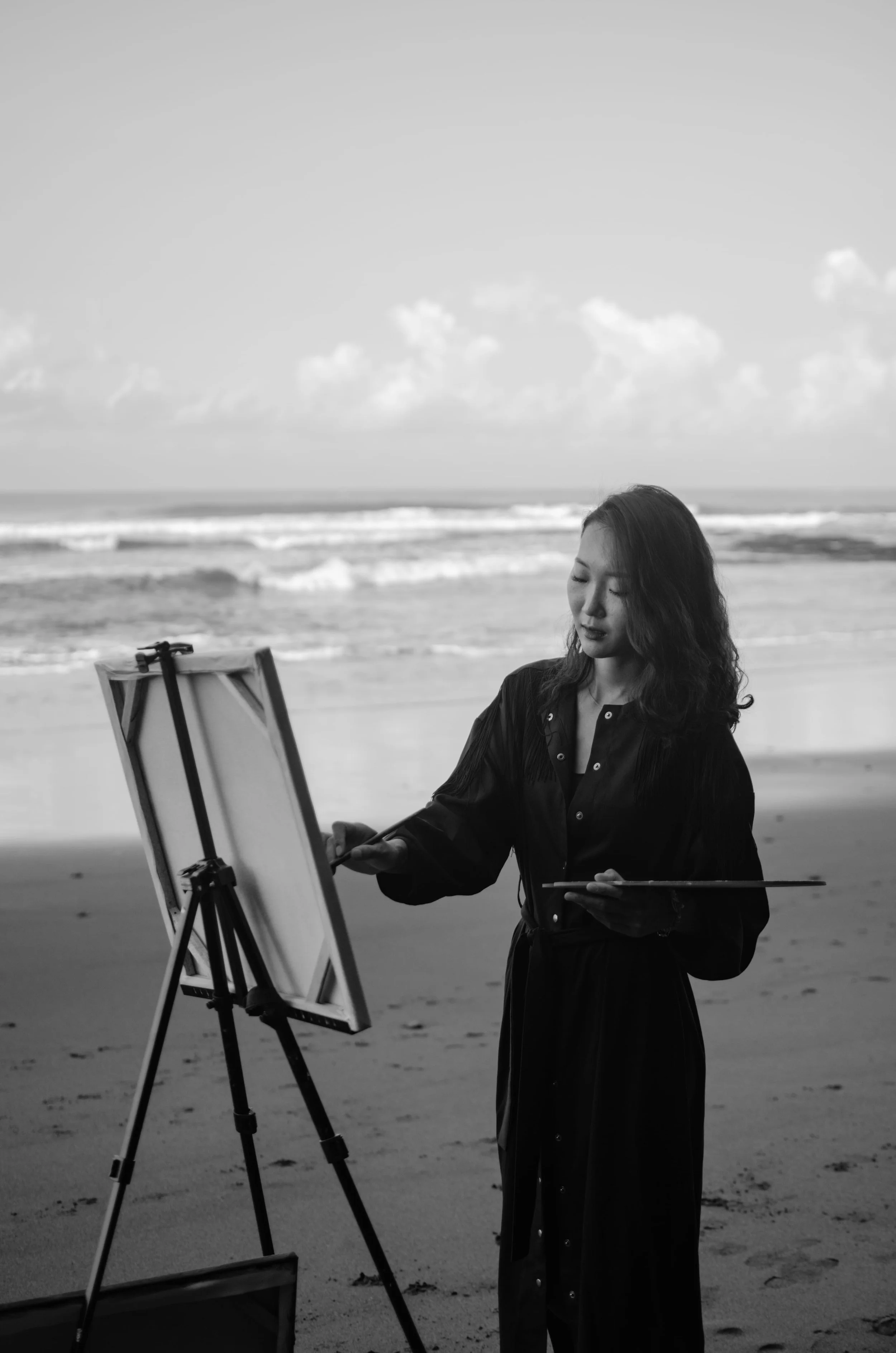 Marina Ogai painting at an easel on the beach, ocean waves behind her, holding palette and brush, black-and-white portrait