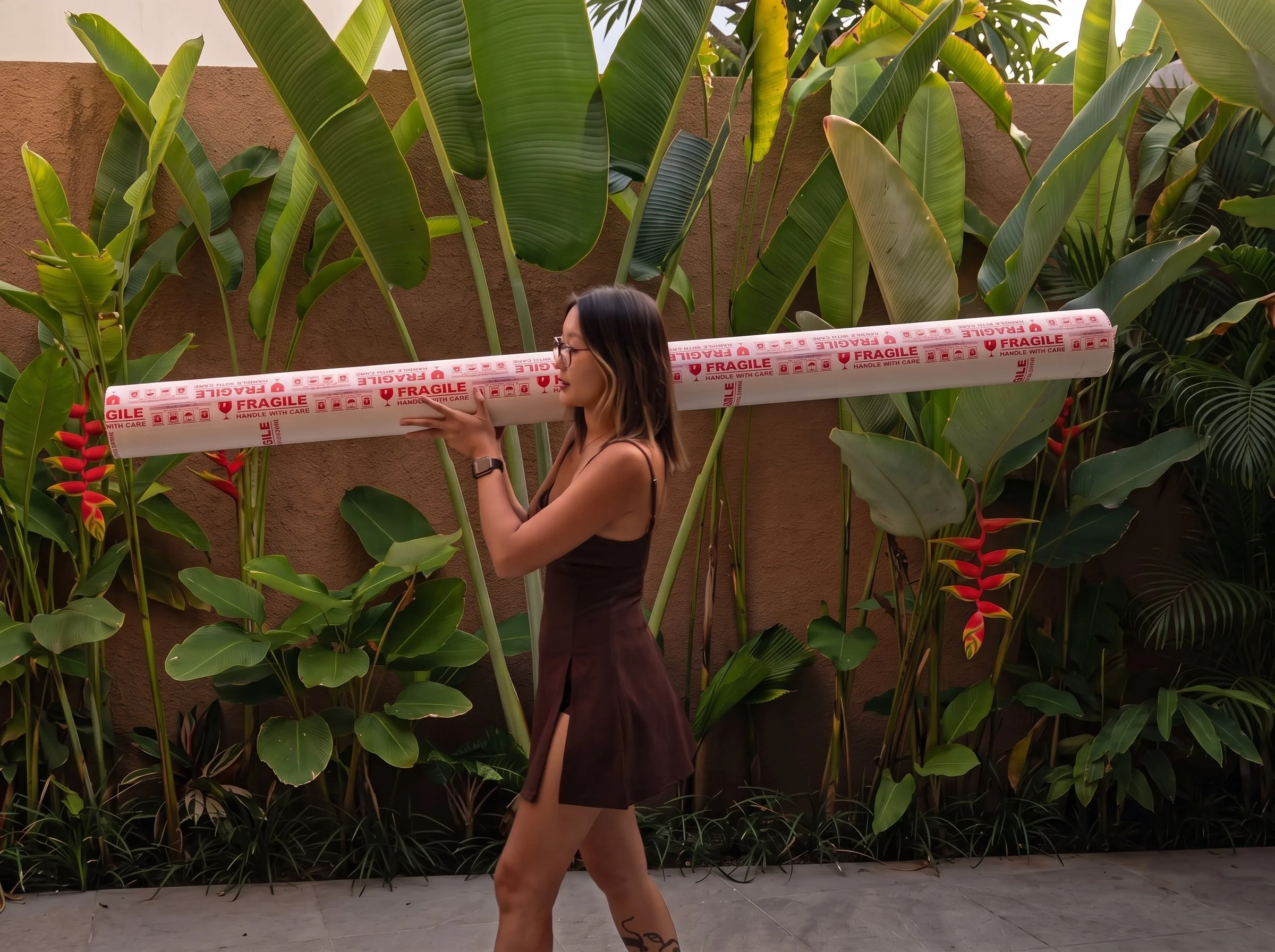 Marina Ogai carrying a rolled oil painting in a long shipping tube marked "Fragile" past tropical plants.