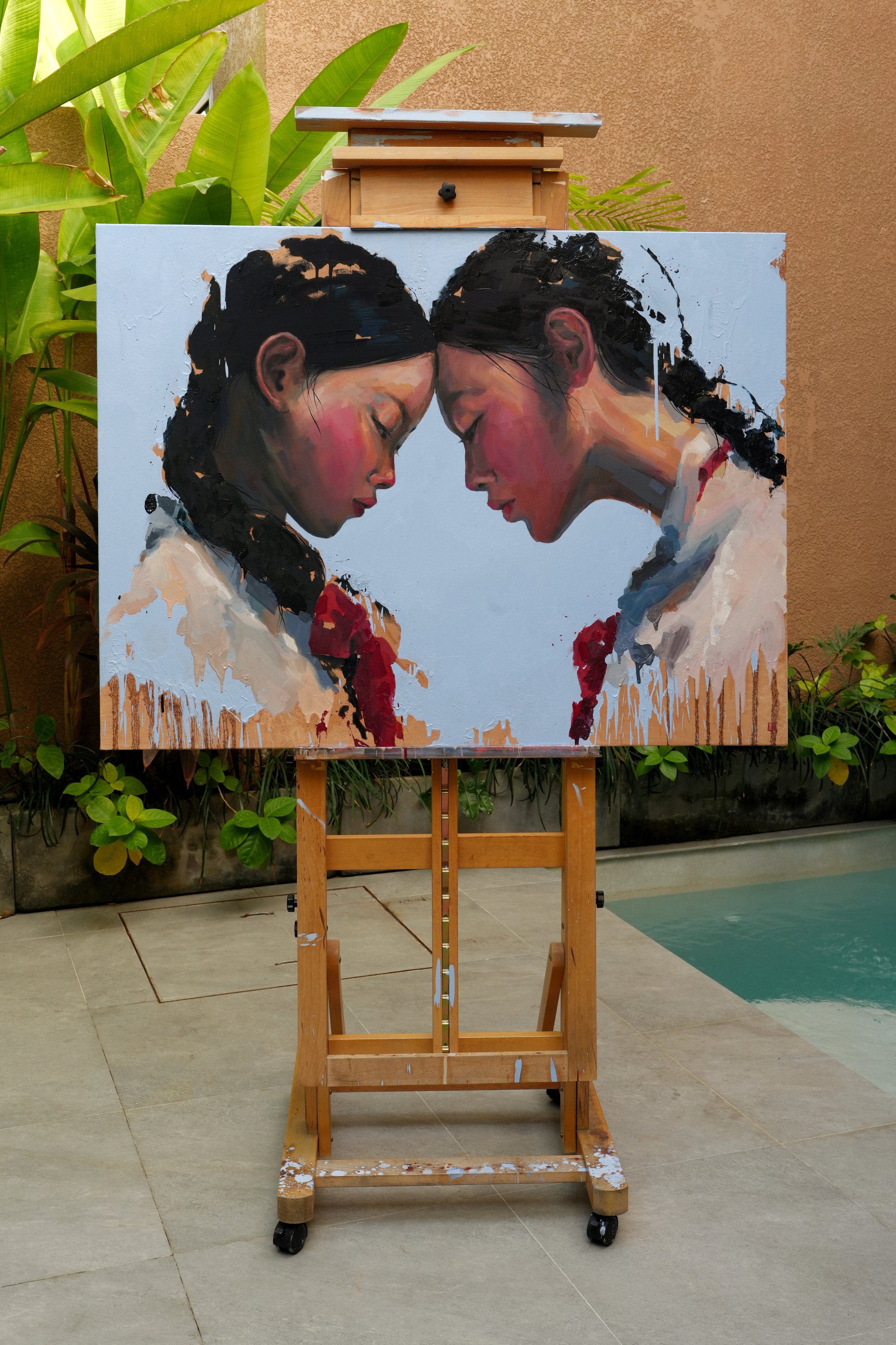 The Encounter by Marina Ogai on a wooden easel outdoors by a pool among tropical plants, showing the scale of the canvas