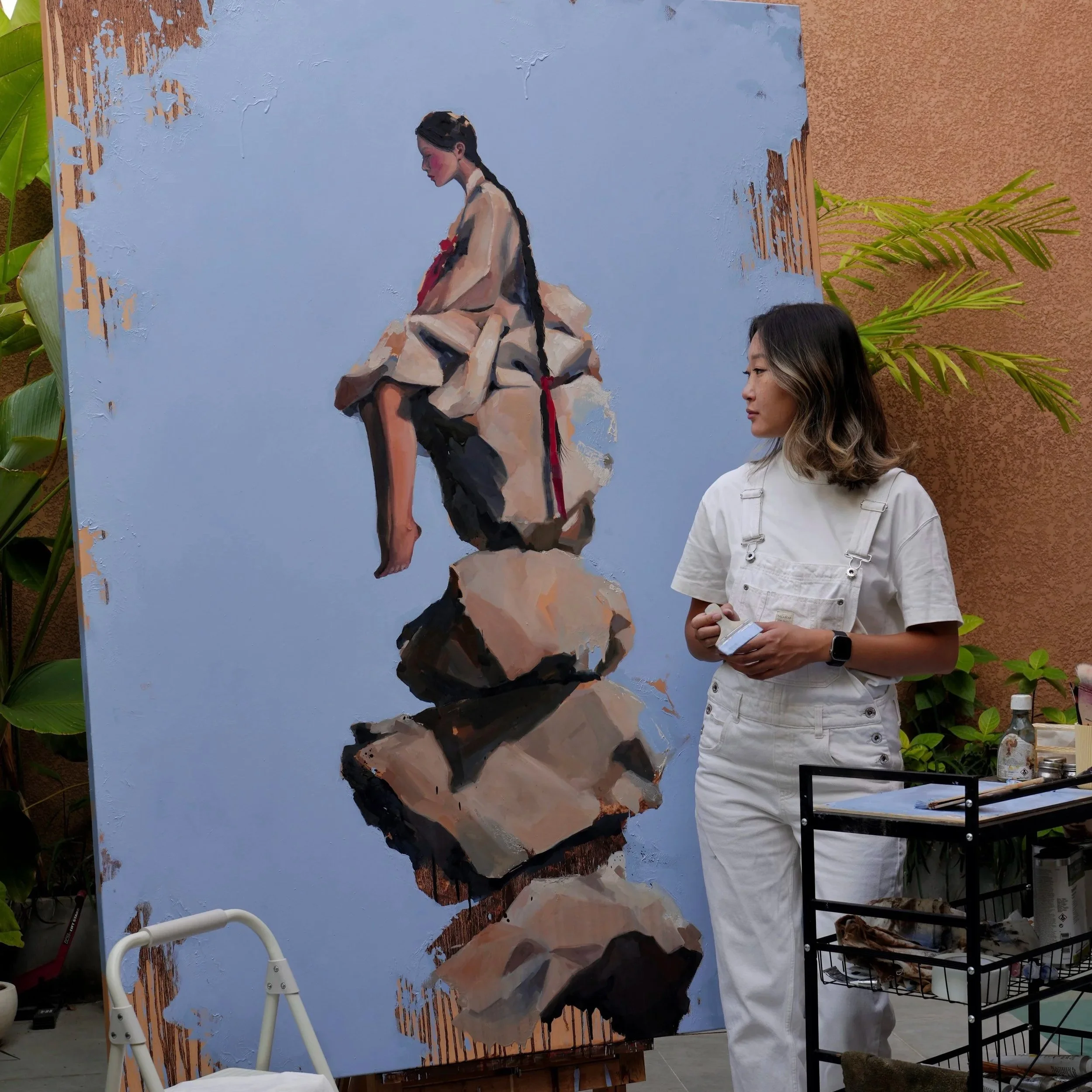 Marina Ogai in her studio next to her large oil on linen painting Cairn, a girl in hanbok sitting on a tall stack of stones