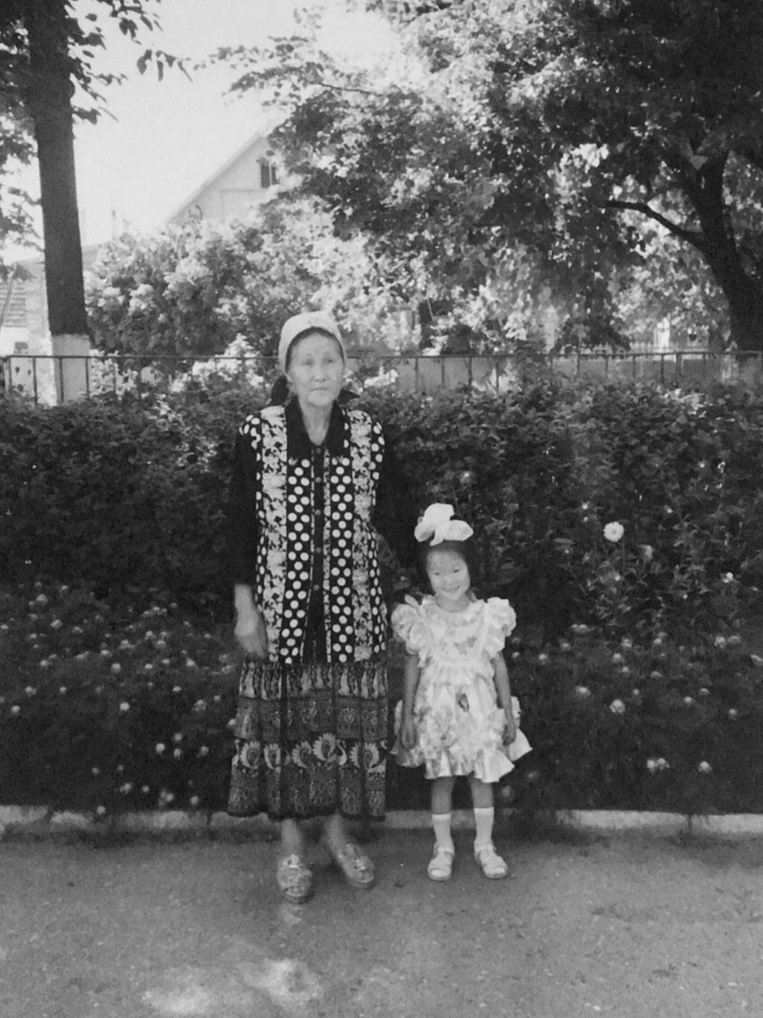 Black-and-white family photo: Marina Ogai as a small girl in a white dress, standing beside her grandmother in a garden