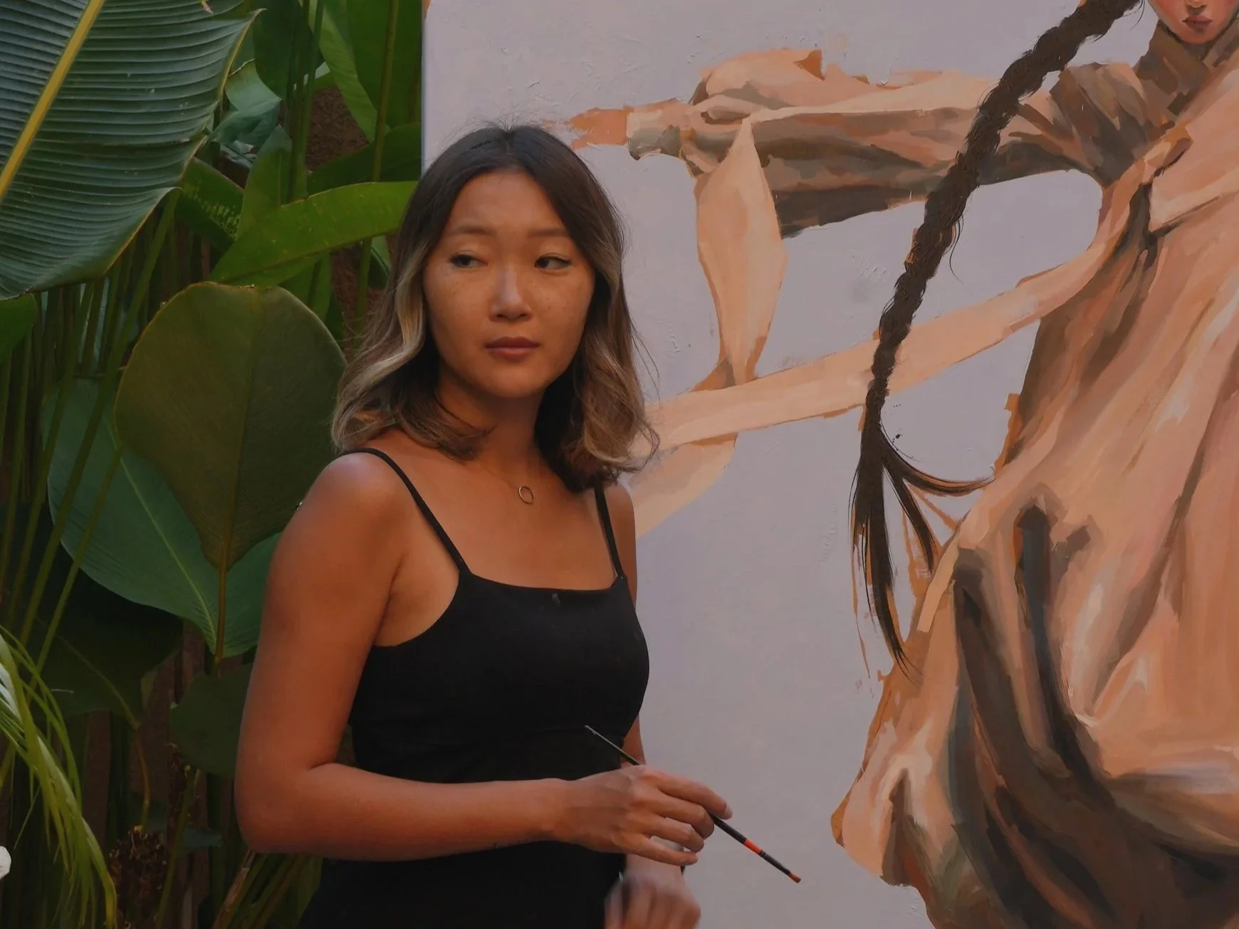 Artist Marina Ogai in her Bali studio with a brush, beside a large oil painting of a woman in hanbok with a long braid.