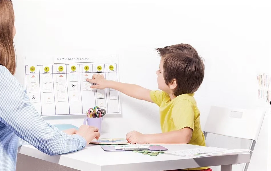 A young boy in a yellow shirt points at a weekly calendar on the wall during a discussion with an adult woman, who is seated at a desk.