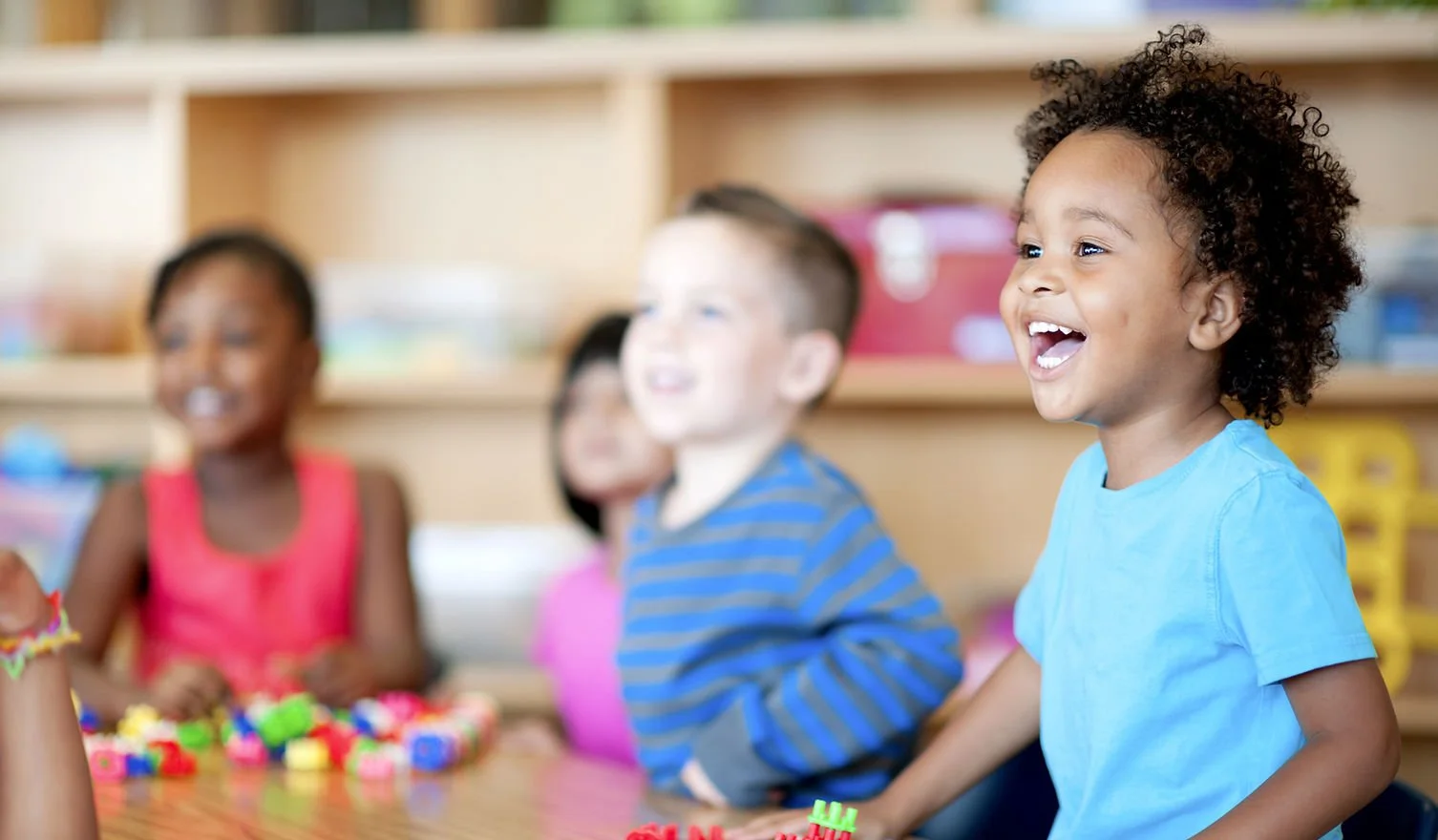 Children sitting at a table, smiling and laughing in a classroom setting.