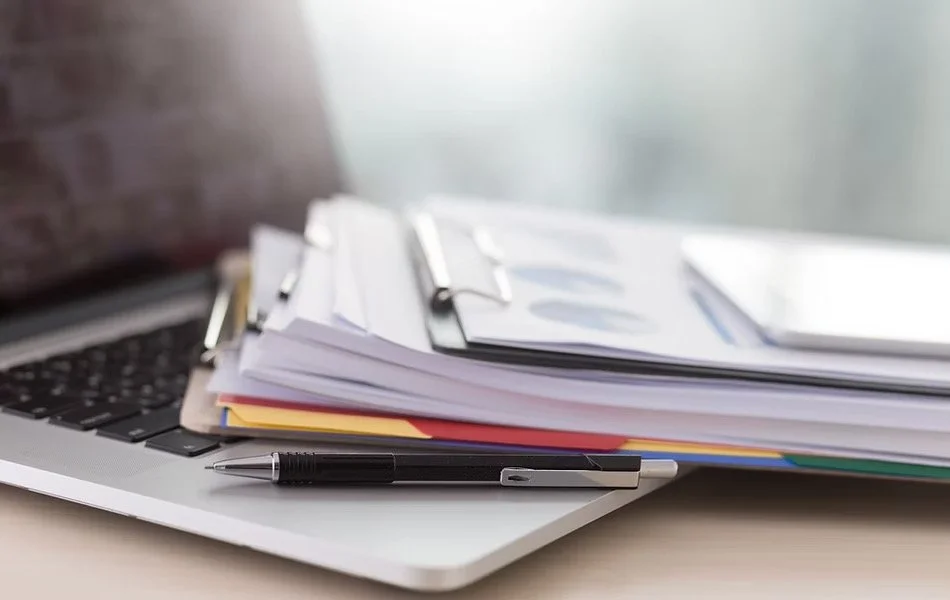 A laptop with a stack of papers on it, a pen placed on the side.