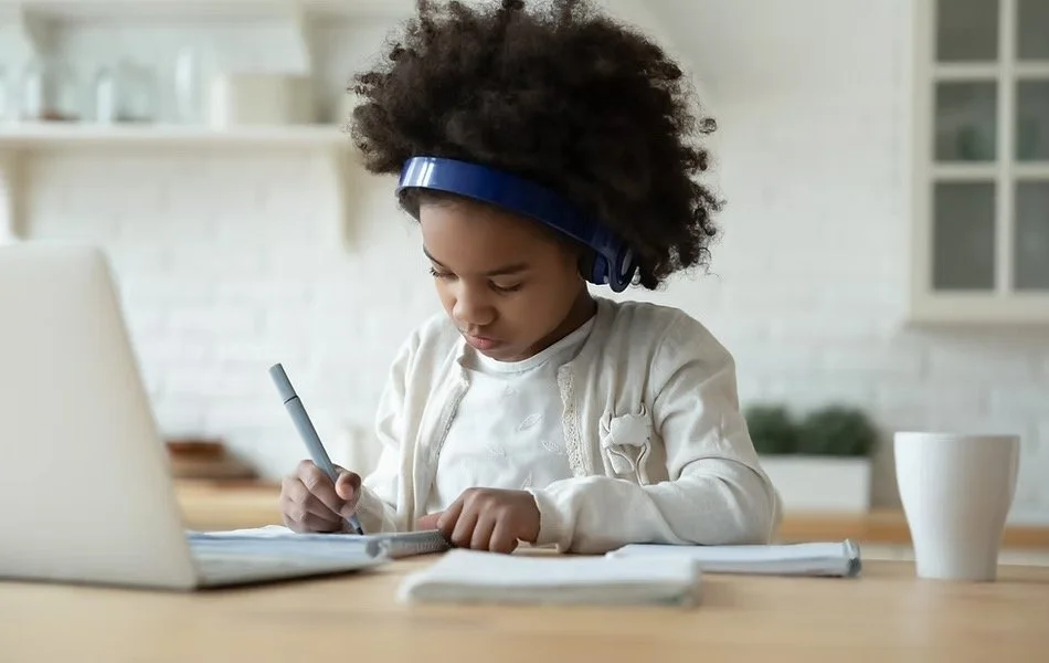 Little girl with afro hairstyle wearing headphones, writing in a notebook at a desk with a laptop and a cup in a bright kitchen.