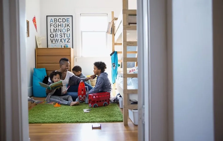 A group of children playing and reading in a brightly lit children's bedroom with a bunk bed, a dresser, and a wall poster of the alphabet.