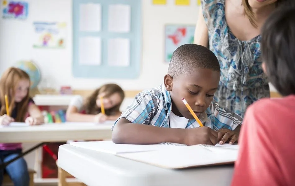 Young boy sitting at desk, writing, in classroom with other children and teacher.