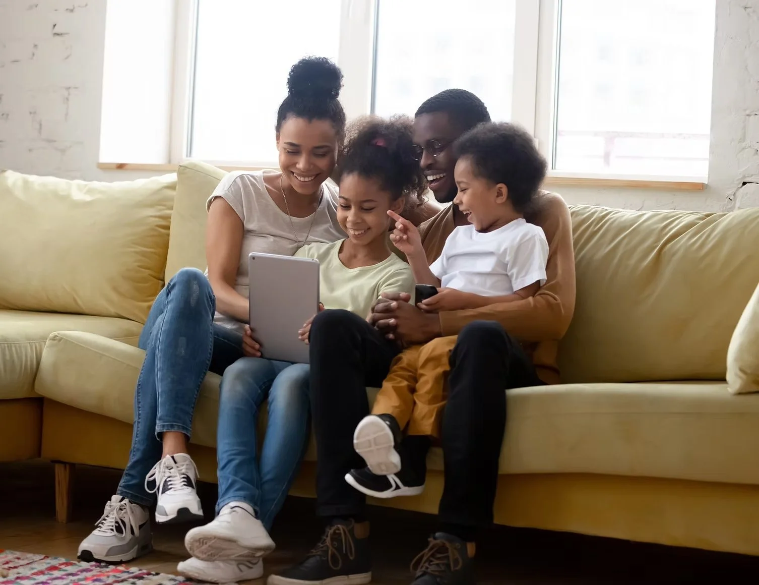 Family of five with dark skin sitting on a yellow couch in a living room, smiling and looking at electronic devices together.