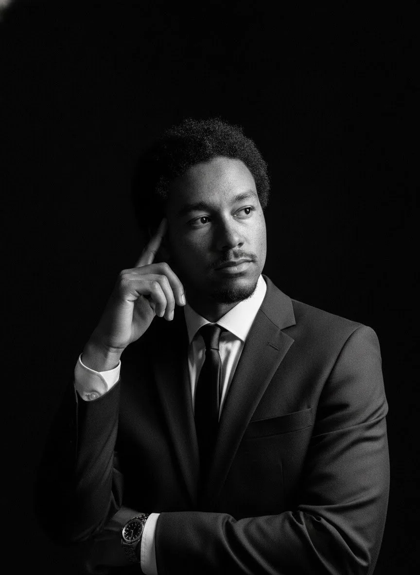 Black and white portrait of a man in a suit, sitting with hand to his temple, looking thoughtfully to the side against a dark background.