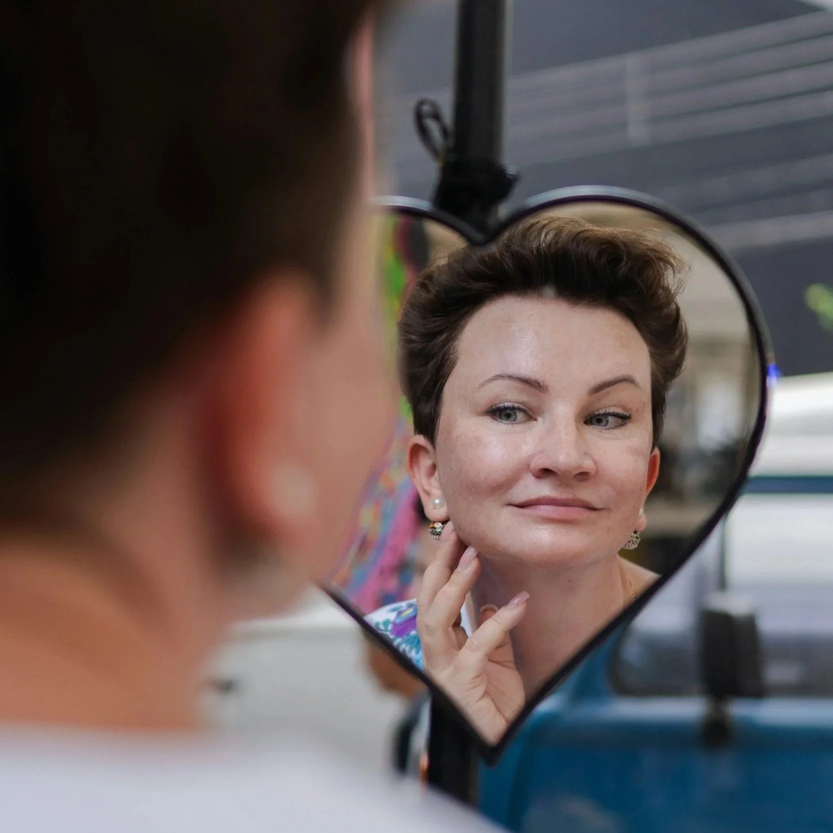 A woman looking at herself in a small mirror, with short brown hair and earrings, examining her own skin