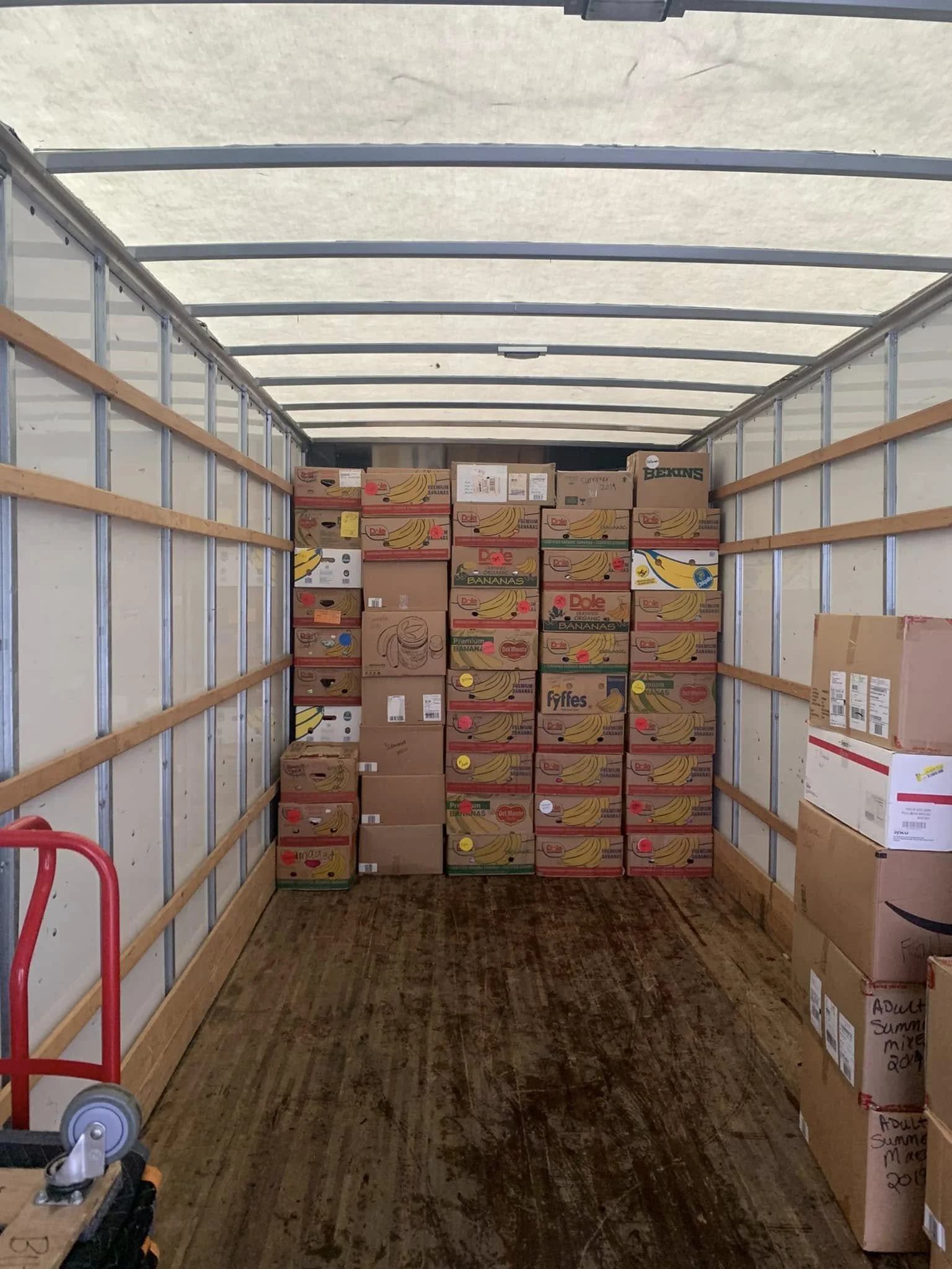 The inside of a moving truck filled with stacked cardboard boxes, mainly labeled with bananas and other produce brands, with a wooden floor and metal walls.