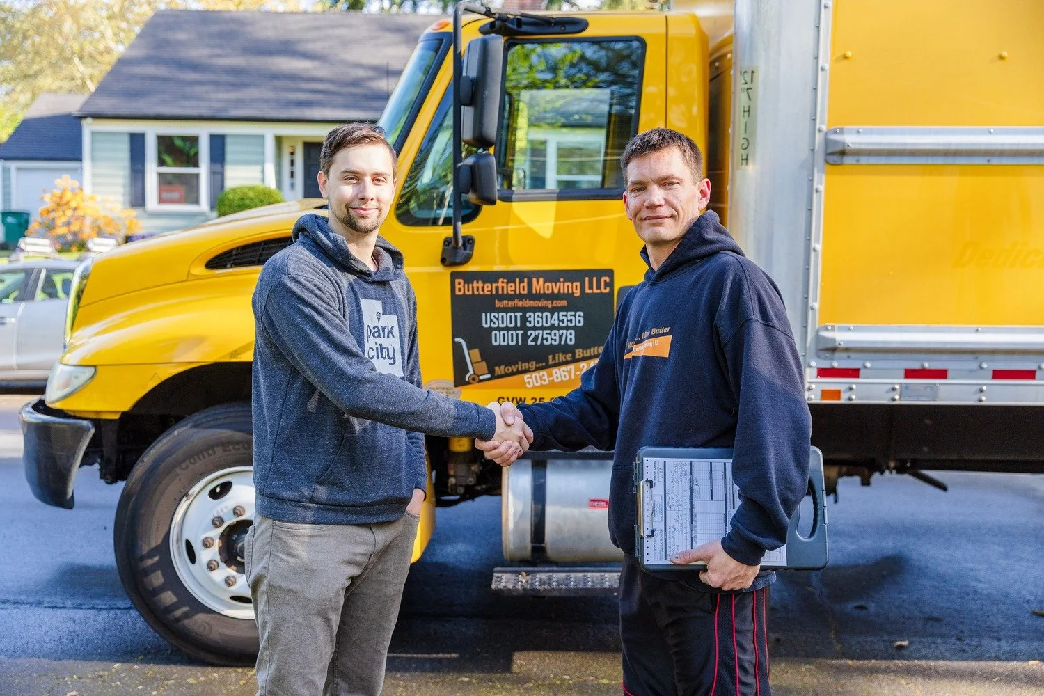 Two men shaking hands in front of a yellow moving truck, with one holding a clipboard, outside a residential neighborhood.
