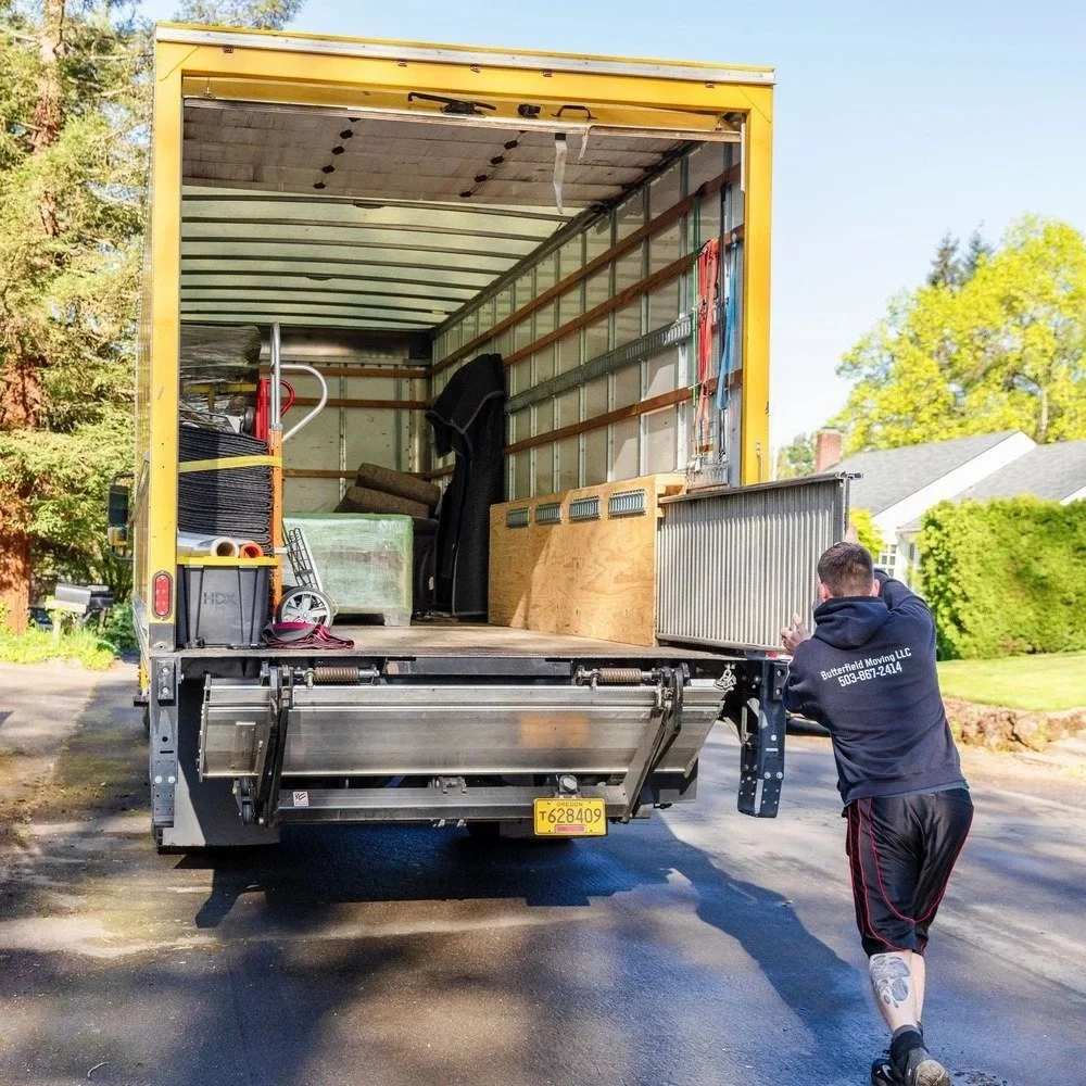 A moving truck with an open back, filled with moving supplies and furniture, parked on a driveway. A person in athletic clothing is standing next to the truck, appearing to be a mover.