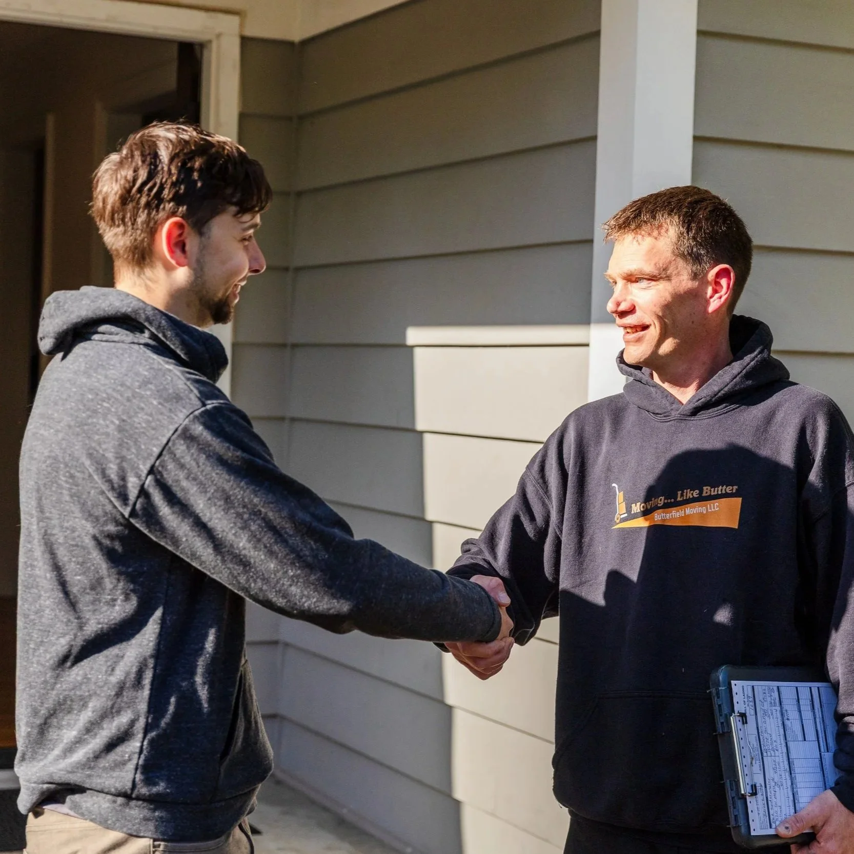 Two men shaking hands outside a house, one holding a clipboard, both smiling.