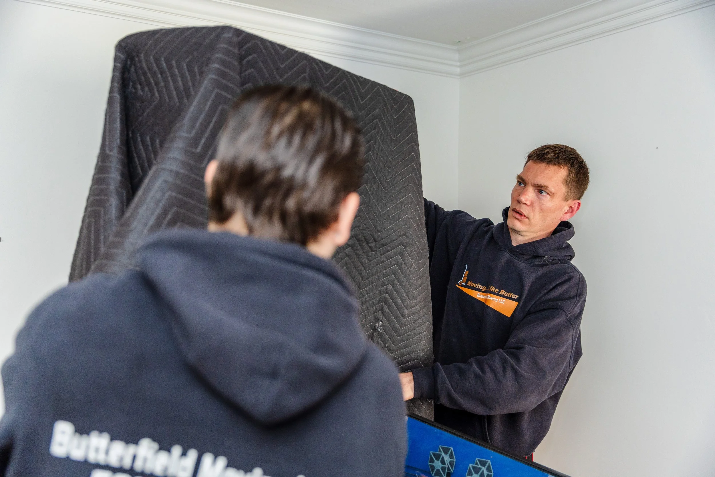 Two men moving a large piece of furniture covered with a quilted moving blanket inside a room with white walls and crown molding. One man appears to be guiding or supporting the furniture, while the other is partially visible in the foreground.