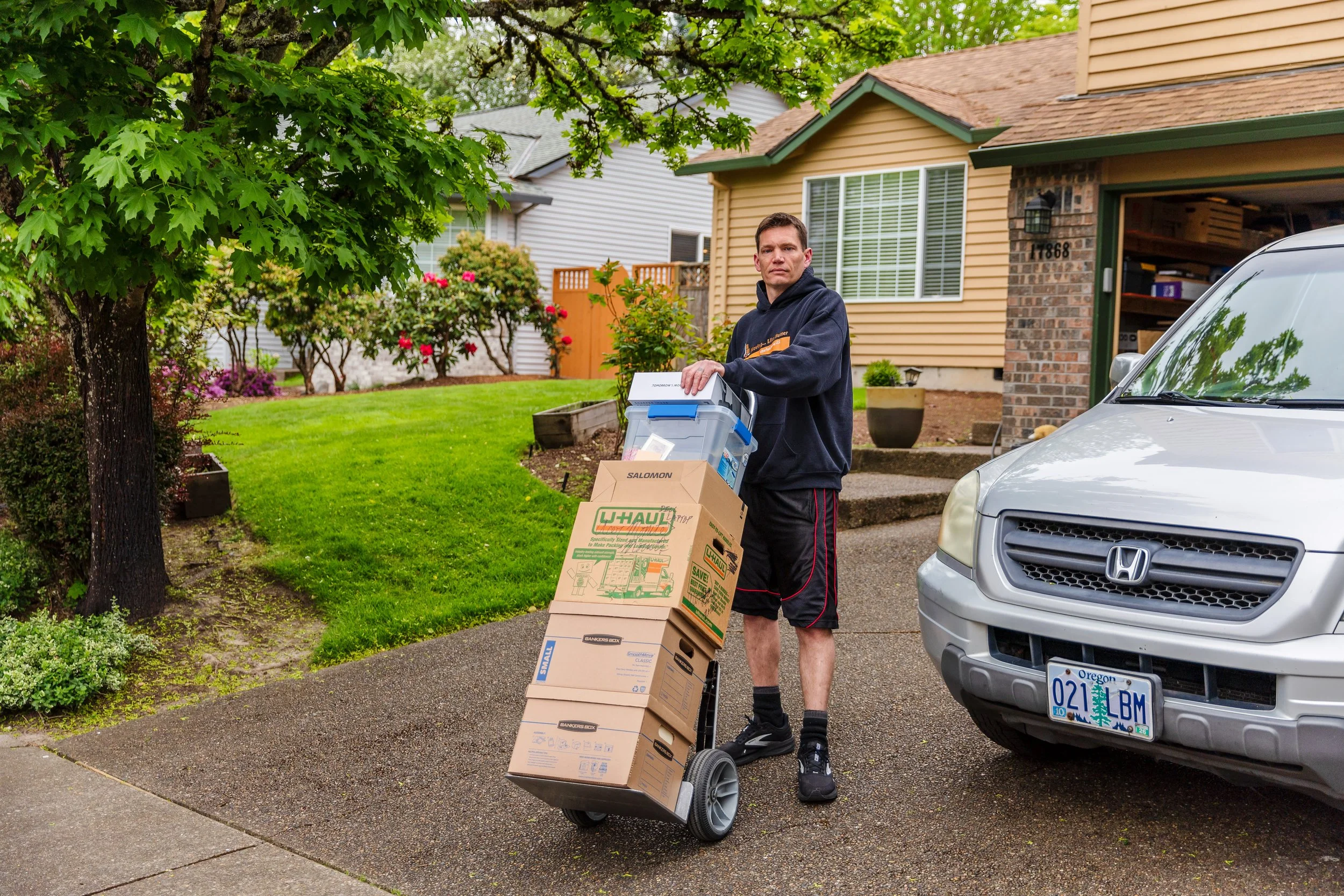 A man in a black hoodie and shorts is delivering in a suburban neighborhood, using a hand truck and carrying several cardboard boxes. There are houses, trees, and a silver car with Oregon license plates in the background.