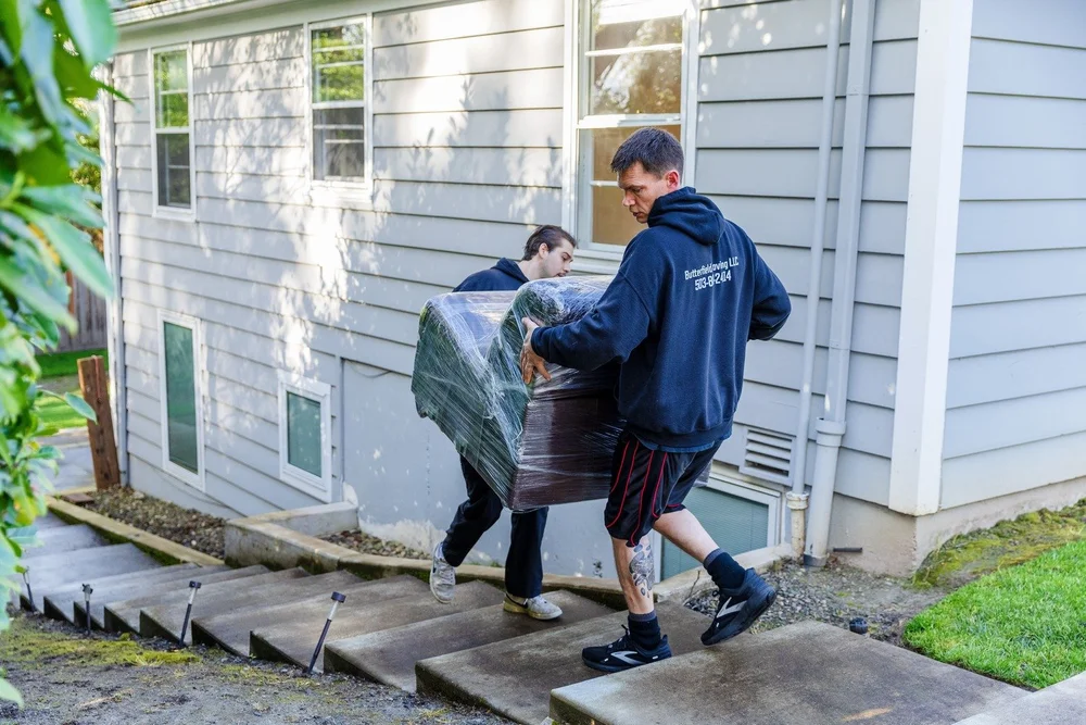 Two movers carrying a wrapped piece down concrete steps beside a house, move in Keizer