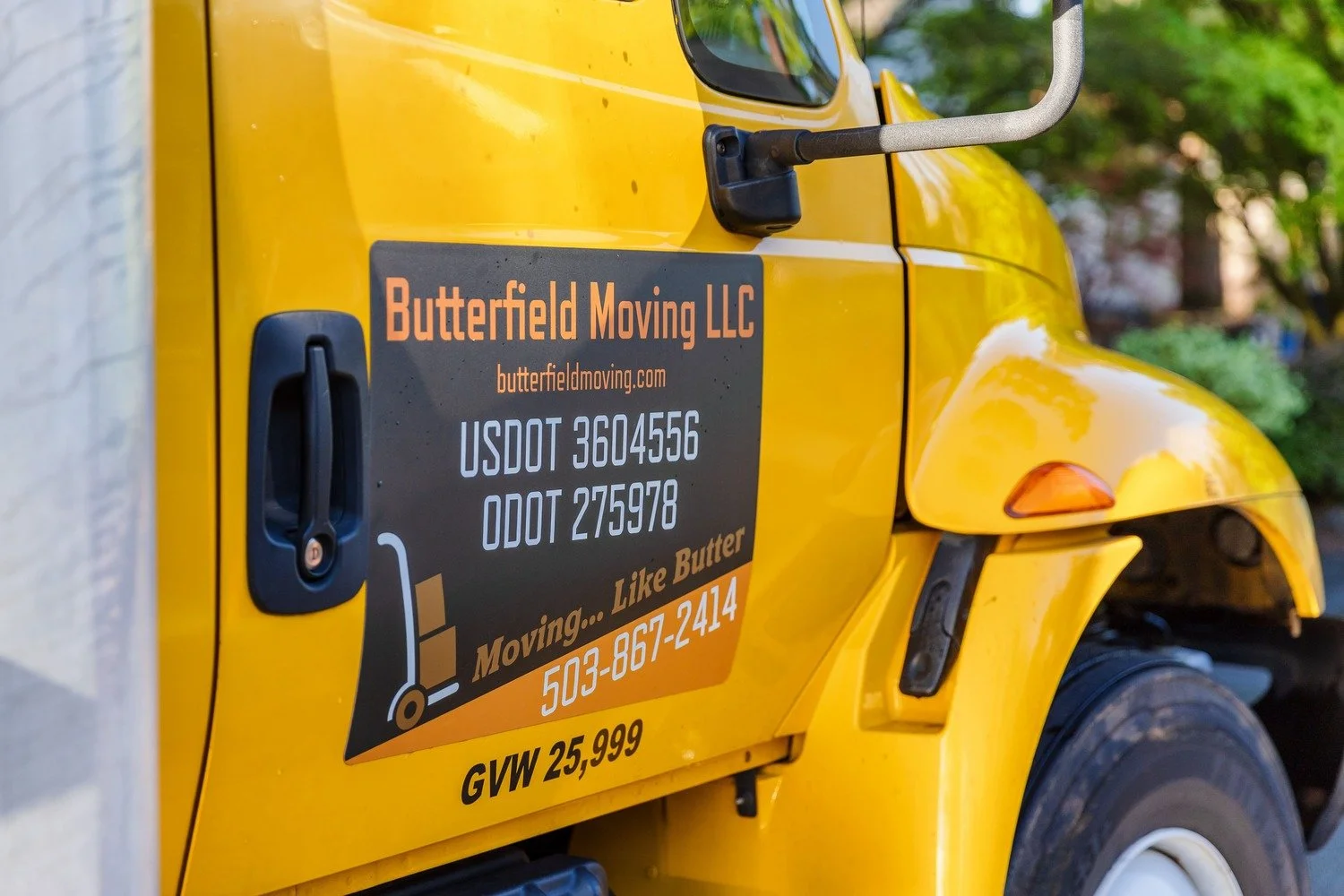 Close-up of a yellow moving truck with a black and orange sign displaying company name 'Butterfield Moving LLC', website, ready-to-move slogan, contact number, and weight capacity, parked outdoors with trees in the background.