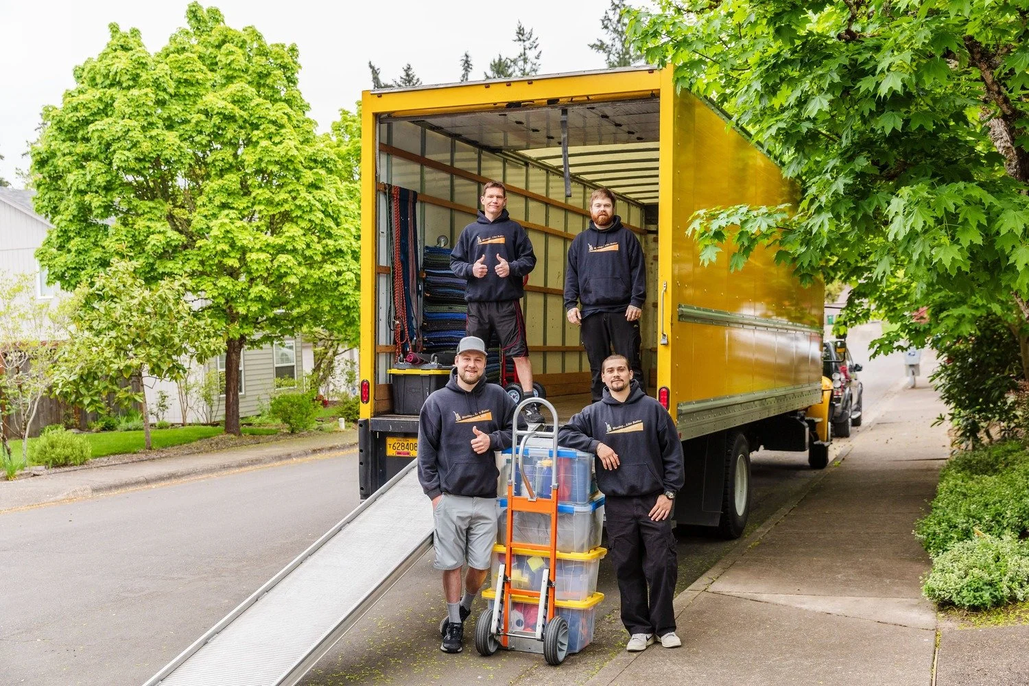 Five men, members of moving crew, standing on and around a yellow moving truck, with some gear and boxes, on a residential street with green trees and houses in the background.