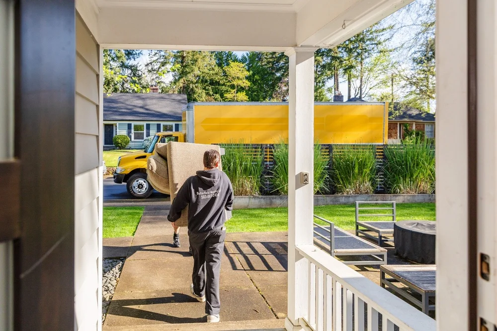 Mover walking a box down a porch path toward the yellow truck, move in St. Helens