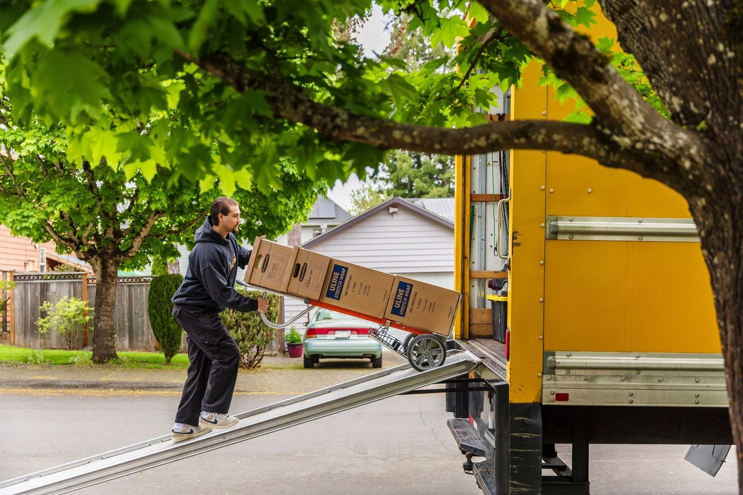 A man loading boxes into a yellow moving truck using a ramp in a suburban neighborhood with green trees and a parked car.