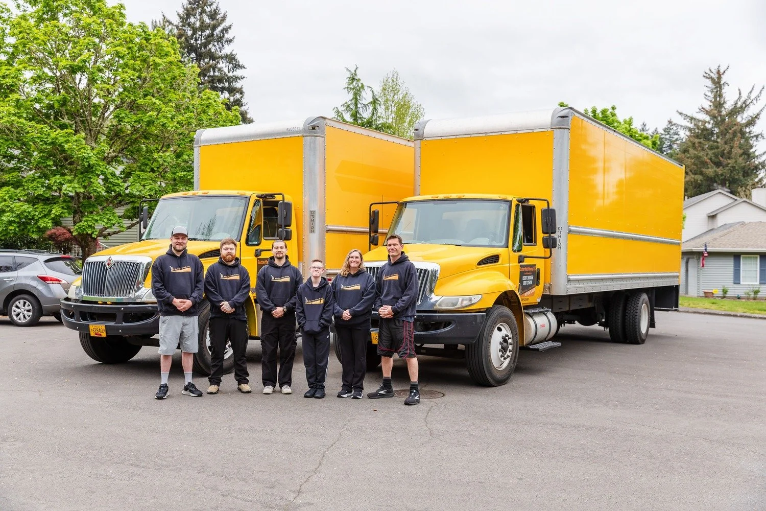 Group of six people standing in front of two large yellow delivery trucks, on a residential street with cars and houses in the background.