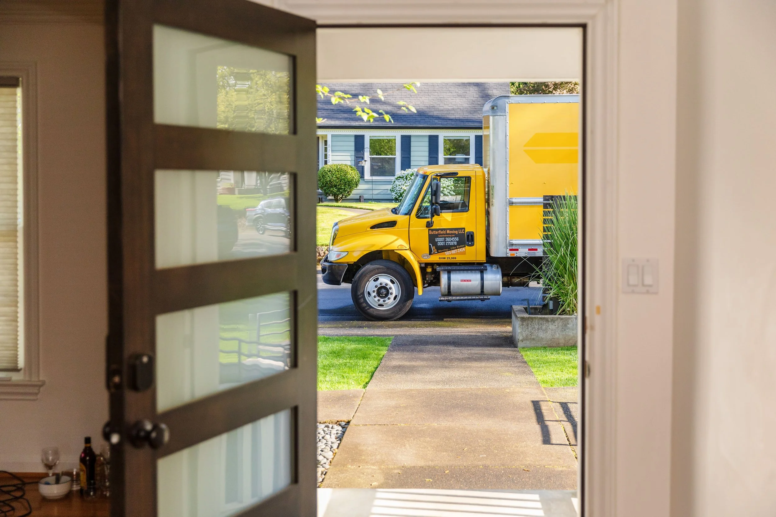 View of a yellow moving truck parked outside a house, seen through an open door with glass panels, with a house and trees in the background.