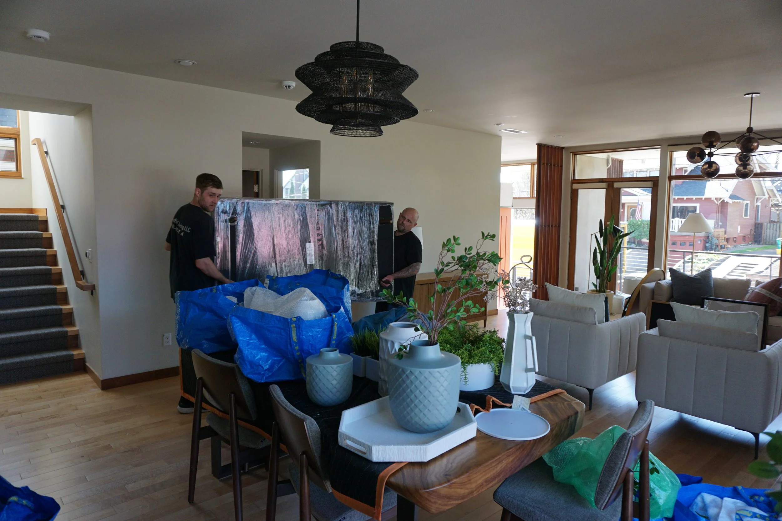 A living room with two men lifting a large glass piece, surrounded by unpacked items and furnishings, with large windows and modern decor.