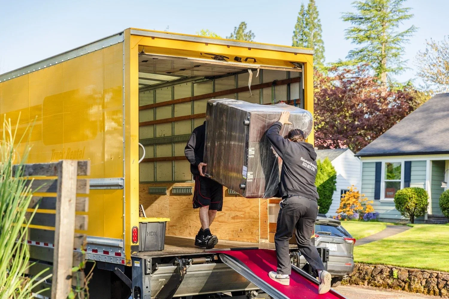 Two men loading a large piece of furniture onto a moving truck with a red ramp in a suburban neighborhood with green lawns and houses.