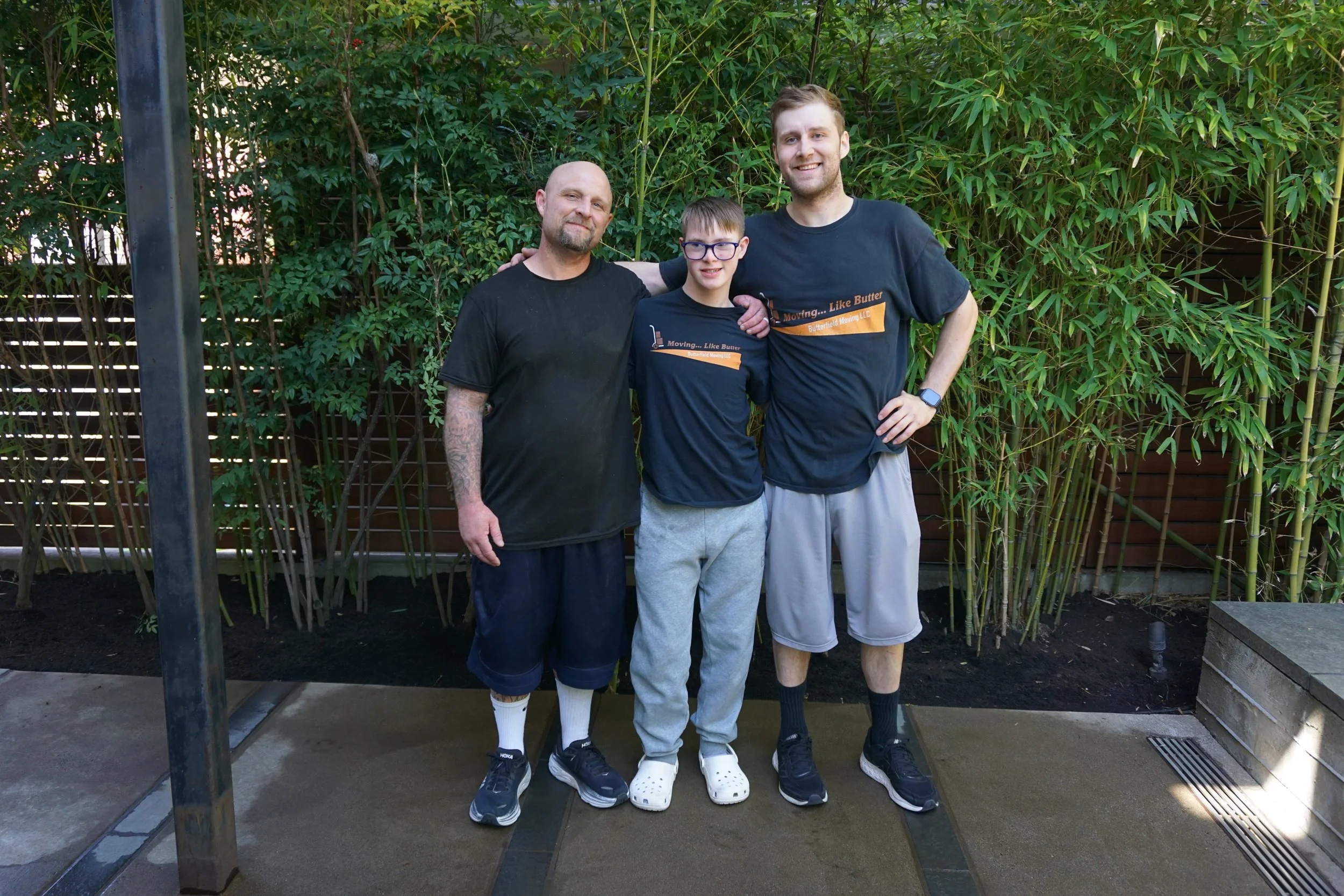 Three males standing together outdoors near green bamboo plants, with a metal post and garden bed in the background. The man on the left has a beard and tattoos on his arm, wearing a black t-shirt and shorts. The boy in the middle has glasses, short hair, and is wearing a black t-shirt with orange text, gray shorts, and Crocs. The man on the right is tall with a beard, wearing a black t-shirt with orange text, gray shorts, and a smartwatch. They are smiling, with two of them having their arms around each other.