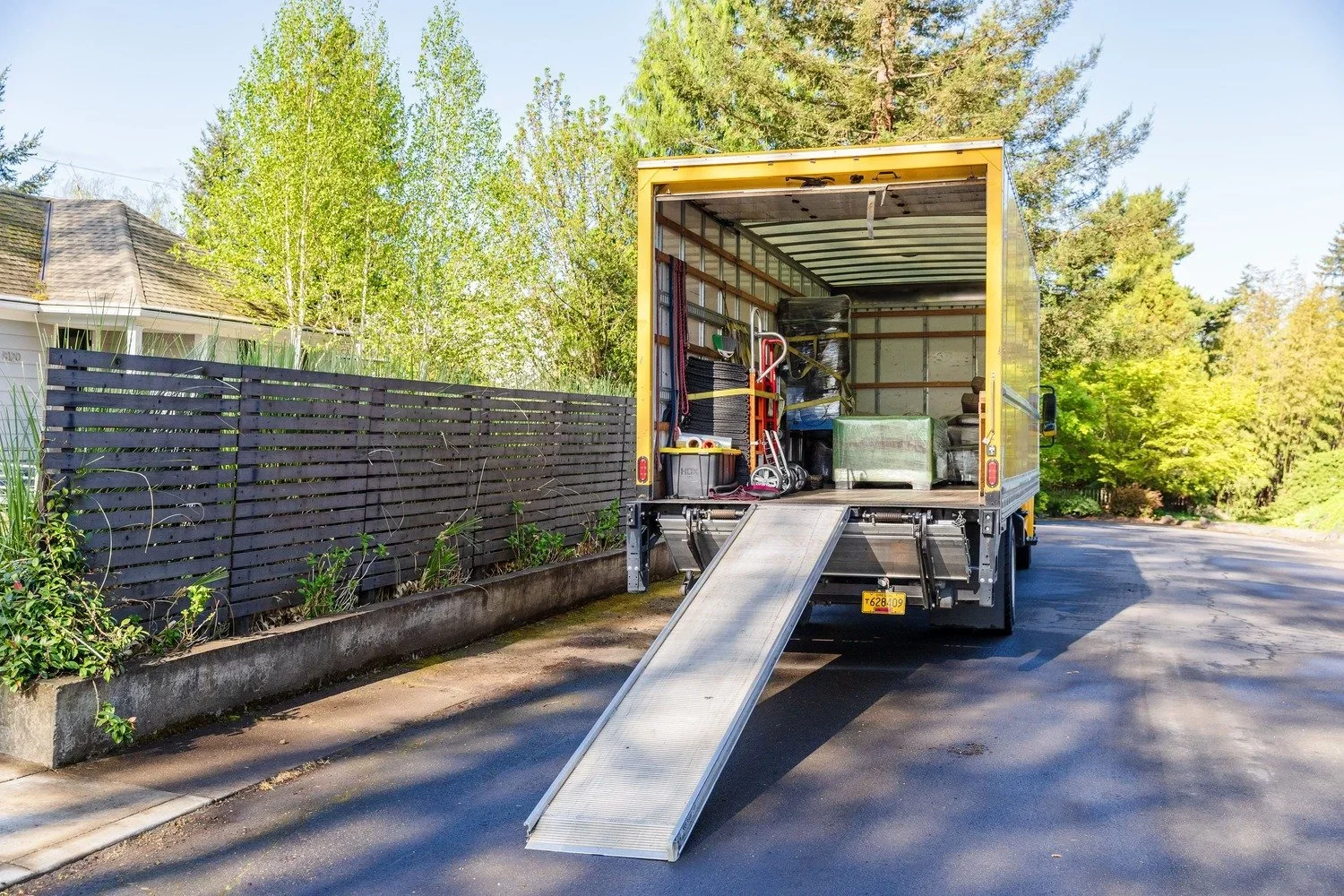 Open yellow moving truck with a ramp extended, parked on a residential street during daytime. The truck contains various household items, including a bicycle, containers, and furniture. Trees and a house with a gray roof are visible in the background.
