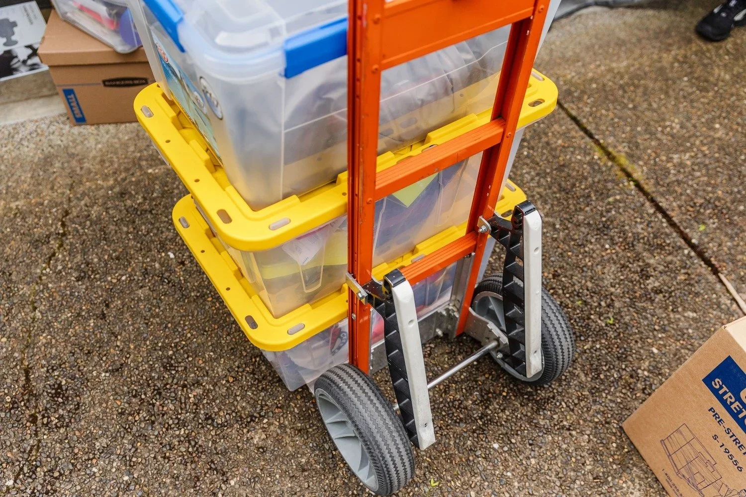A utility cart with yellow and orange plastic bins containing various items, on a rough outdoor surface. A cardboard box with blue print is next to it.