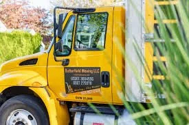 Yellow moving truck with company name and contact information on the door, parked outdoors next to green foliage.