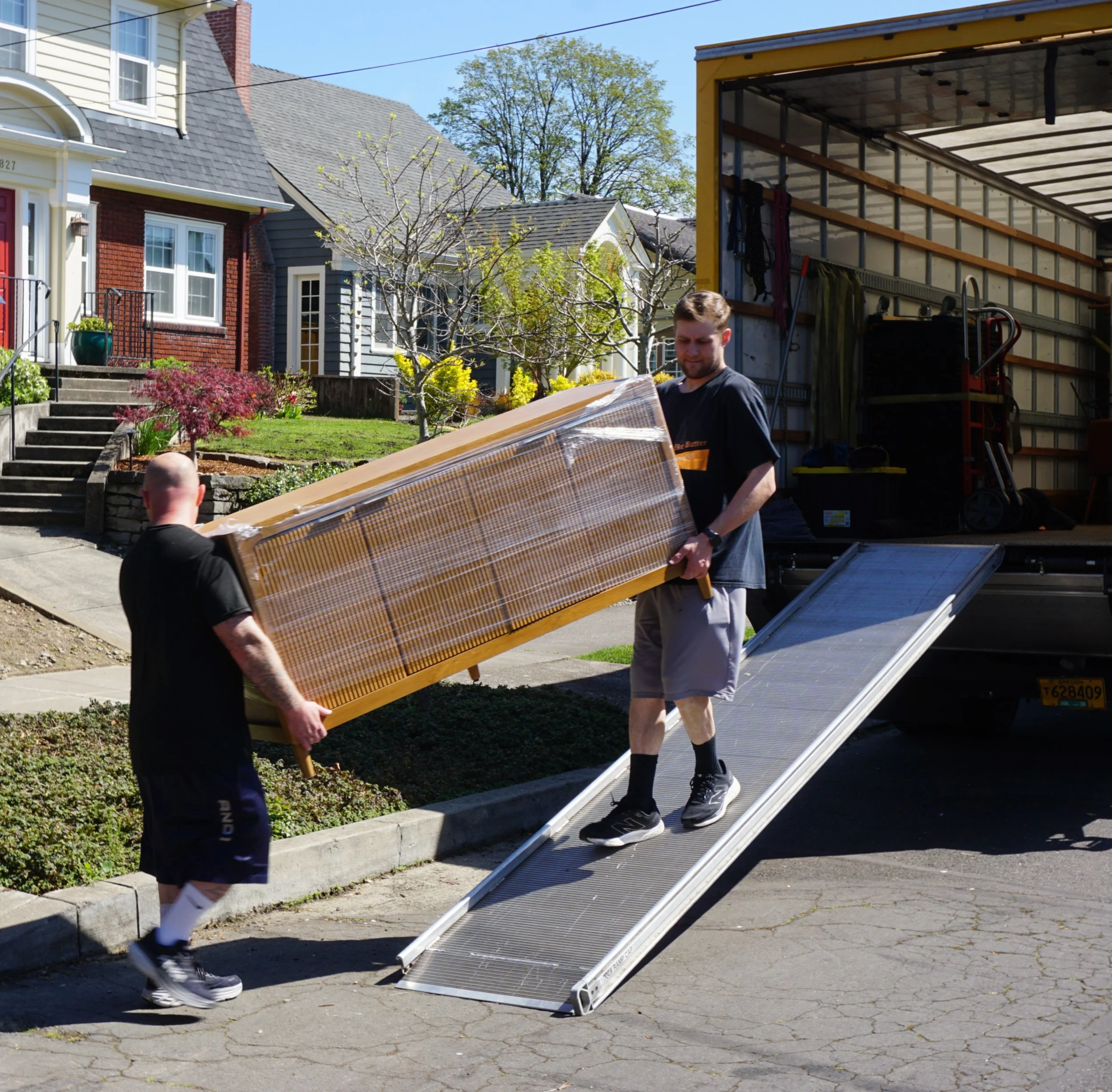 Two men are moving a large glass window screen onto a truck parked on a residential street. One man is lifting the screen while the other helps carry it. The background shows colorful houses, a tree, and a clear blue sky, indicating a sunny day.