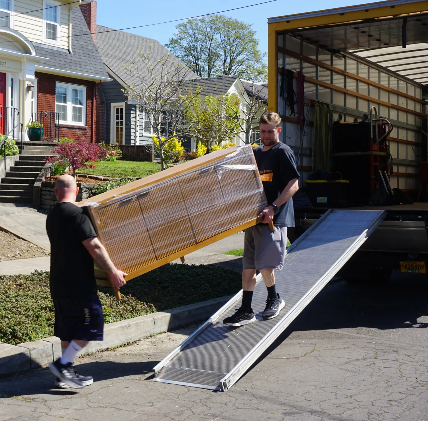 Two movers carrying a wrapped piece up a ramp outside a brick house, move in Newberg