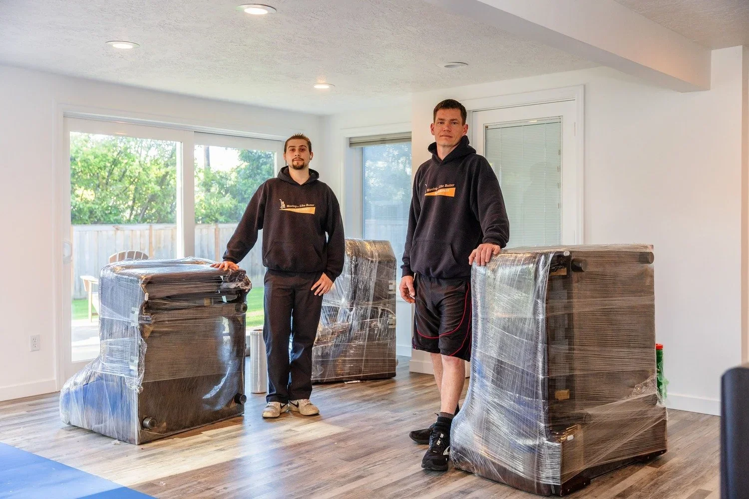 Two movers in black hoodies standing in a living room with wrapped furniture, overlooking a backyard with trees.