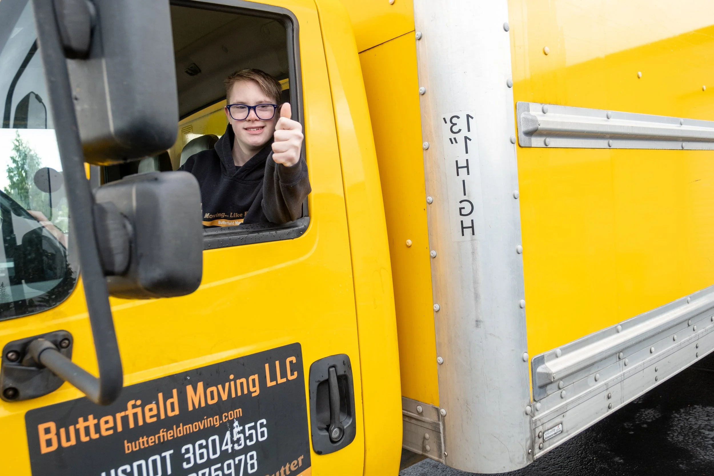 A young man with glasses giving a thumbs-up while sitting in the driver's seat of a bright yellow moving truck.