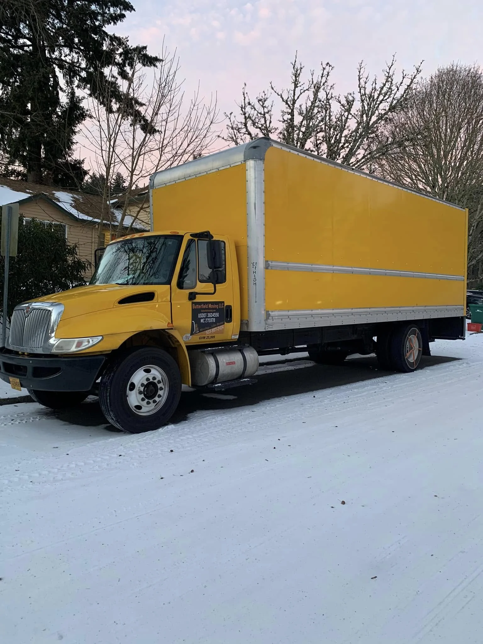A yellow moving truck parked on a snow-covered street in front of a house with bare trees in the background during winter.