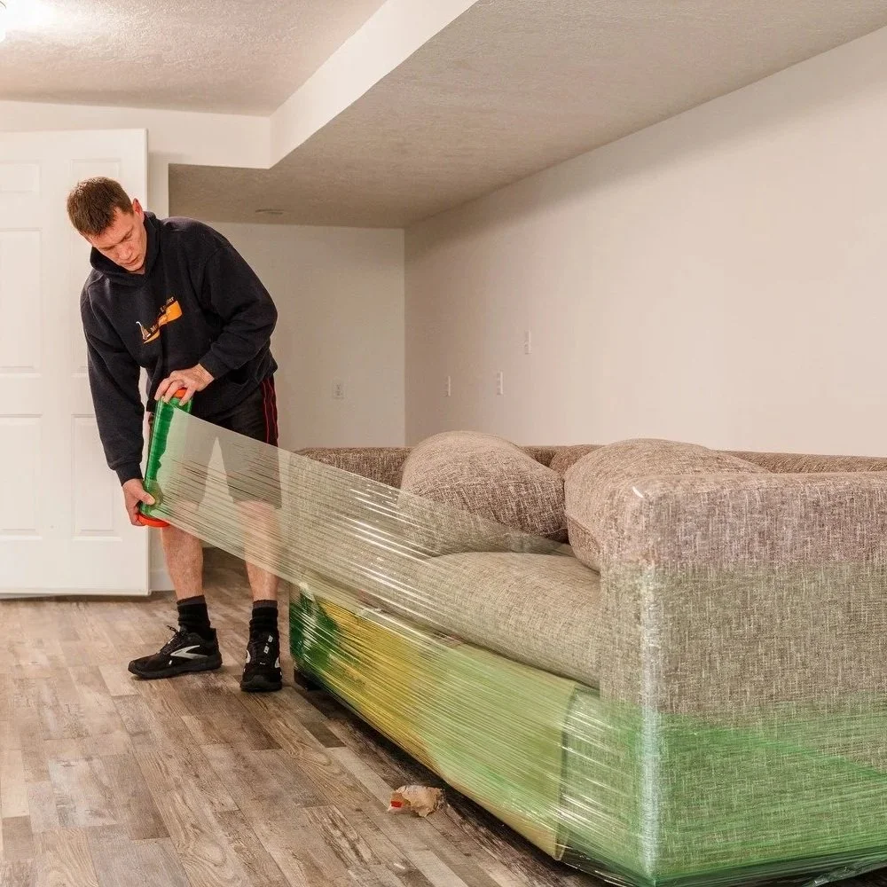 A man is wrapping a couch with plastic wrap in a basement or living room with wooden flooring and white walls.